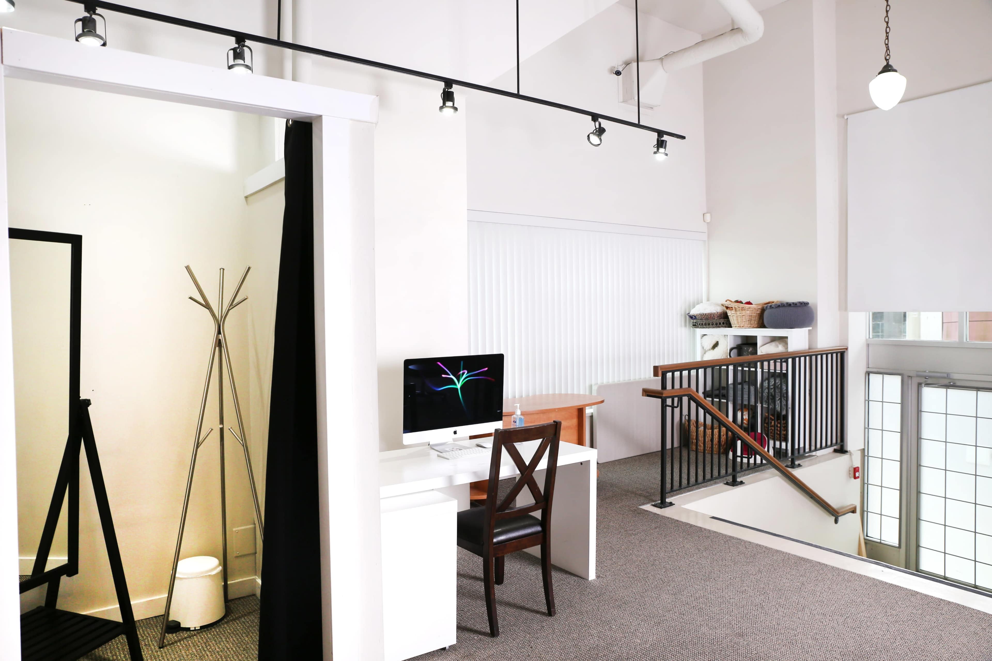 A modern office space featuring a white desk with a computer, a tall mirror, and a staircase leading to a lower level, all within a well-lit, open area.