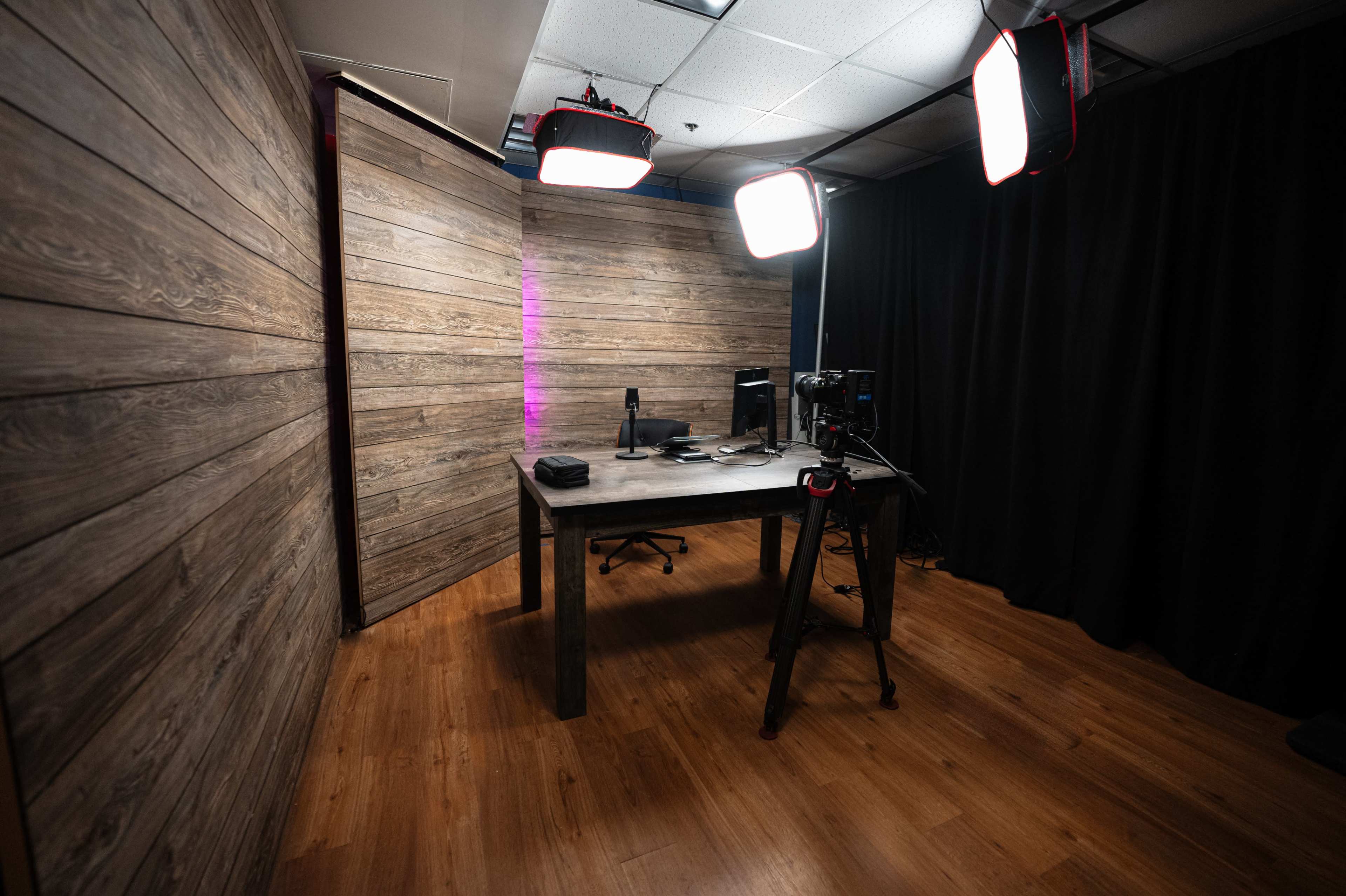 A small, well-lit studio space featuring a wooden table, computer monitor, and camera equipment against a backdrop of wooden walls and a black curtain.