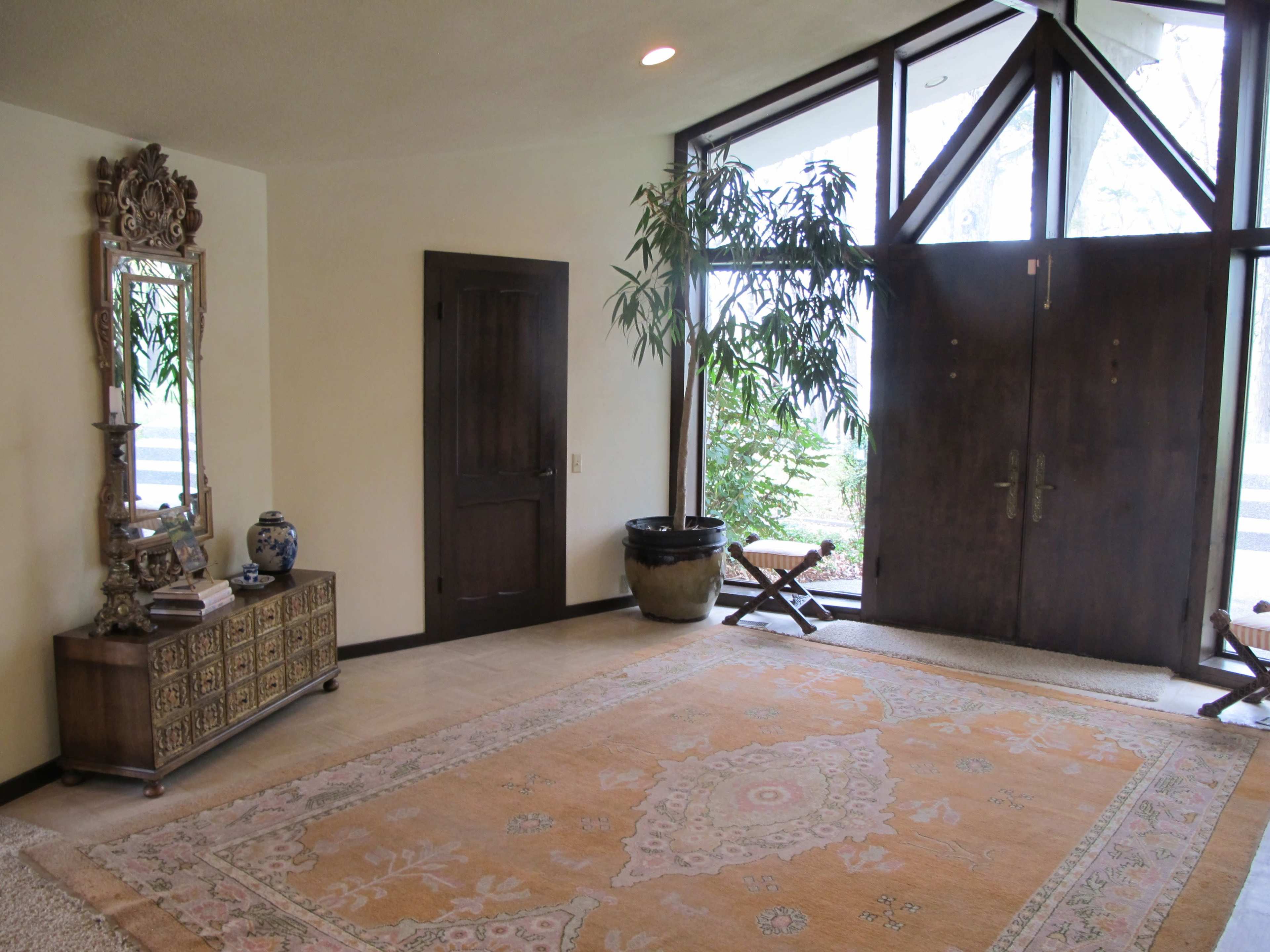The image shows a spacious entryway with a large area rug, a mirror atop a decorative console table, and large windows allowing natural light to enter.