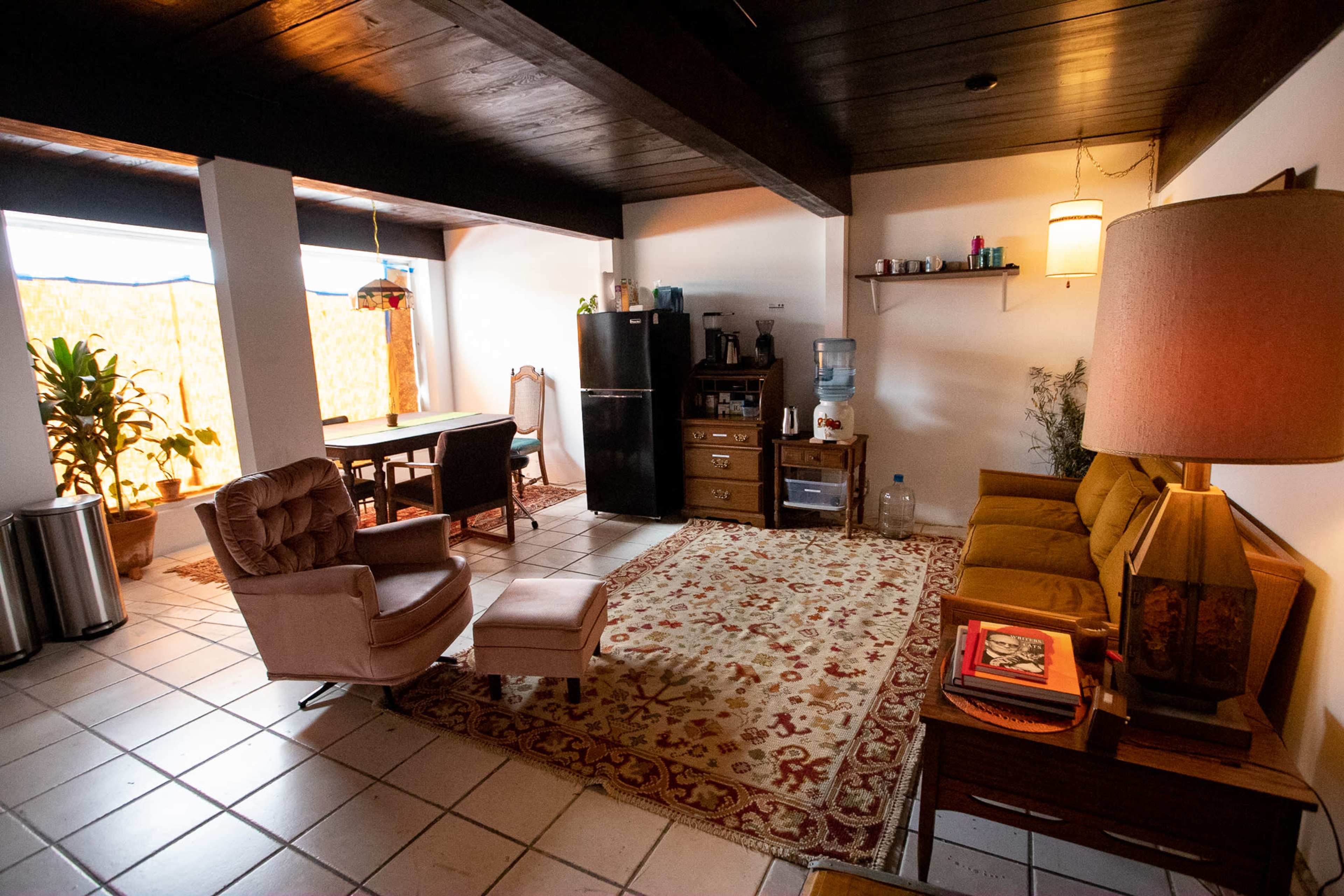 The image shows a cozy living space featuring a seating area with a sofa and armchair, a dining table, and a kitchenette with a refrigerator and water cooler.