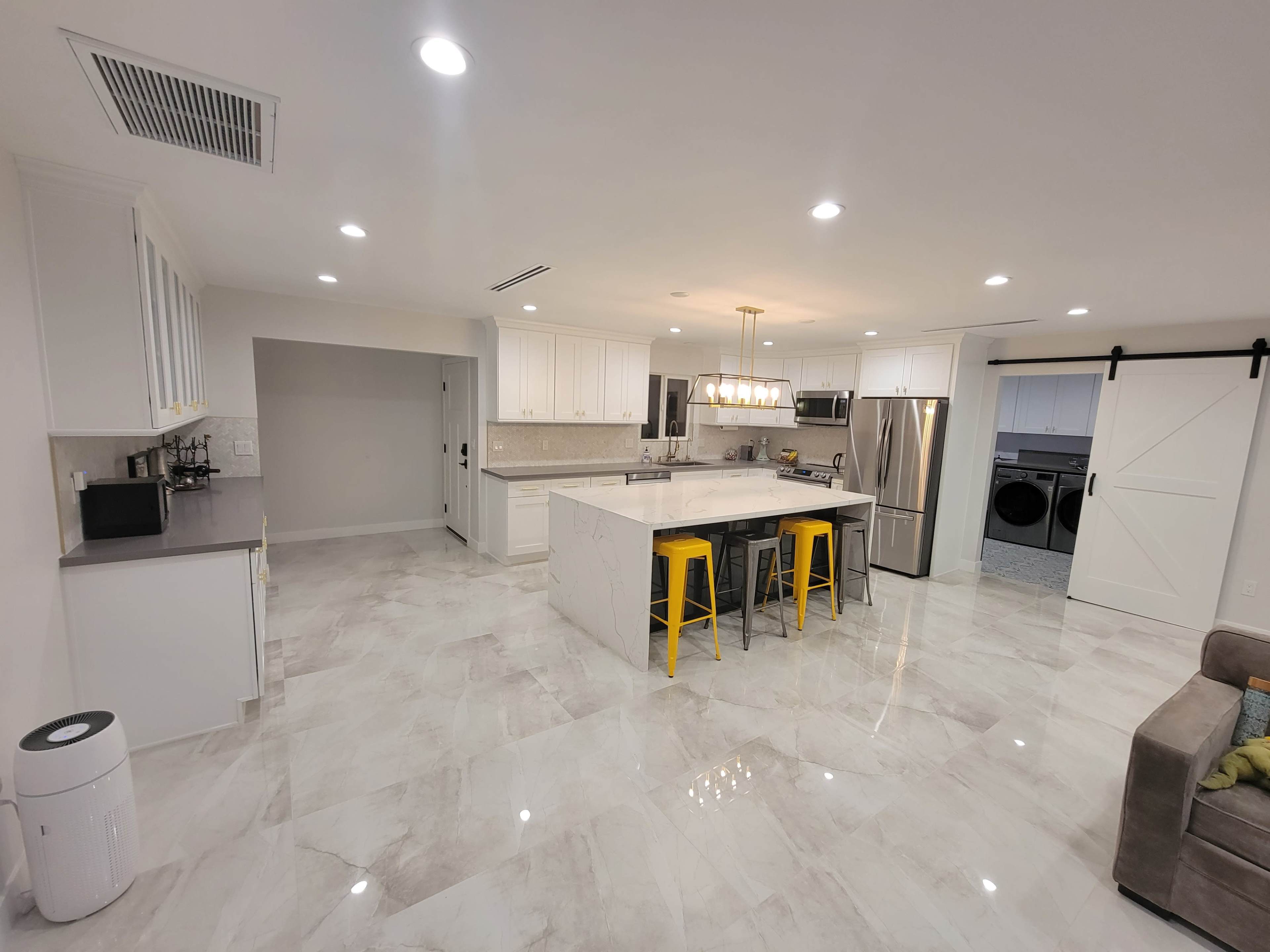 A modern kitchen features white cabinetry, a central island with yellow bar stools, stainless steel appliances, and polished marble flooring.
