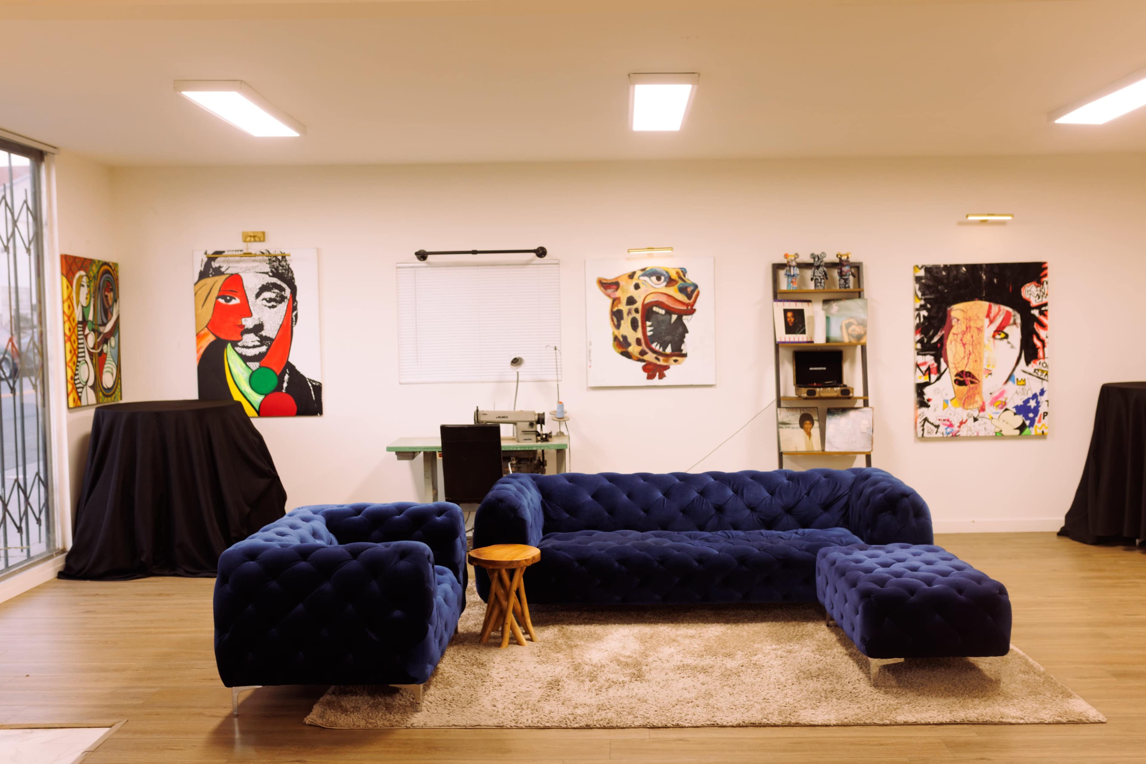 The image shows a spacious room with a blue tufted sofa set, a small wooden stool, and vibrant artwork displayed on the walls, alongside a desk and shelves.