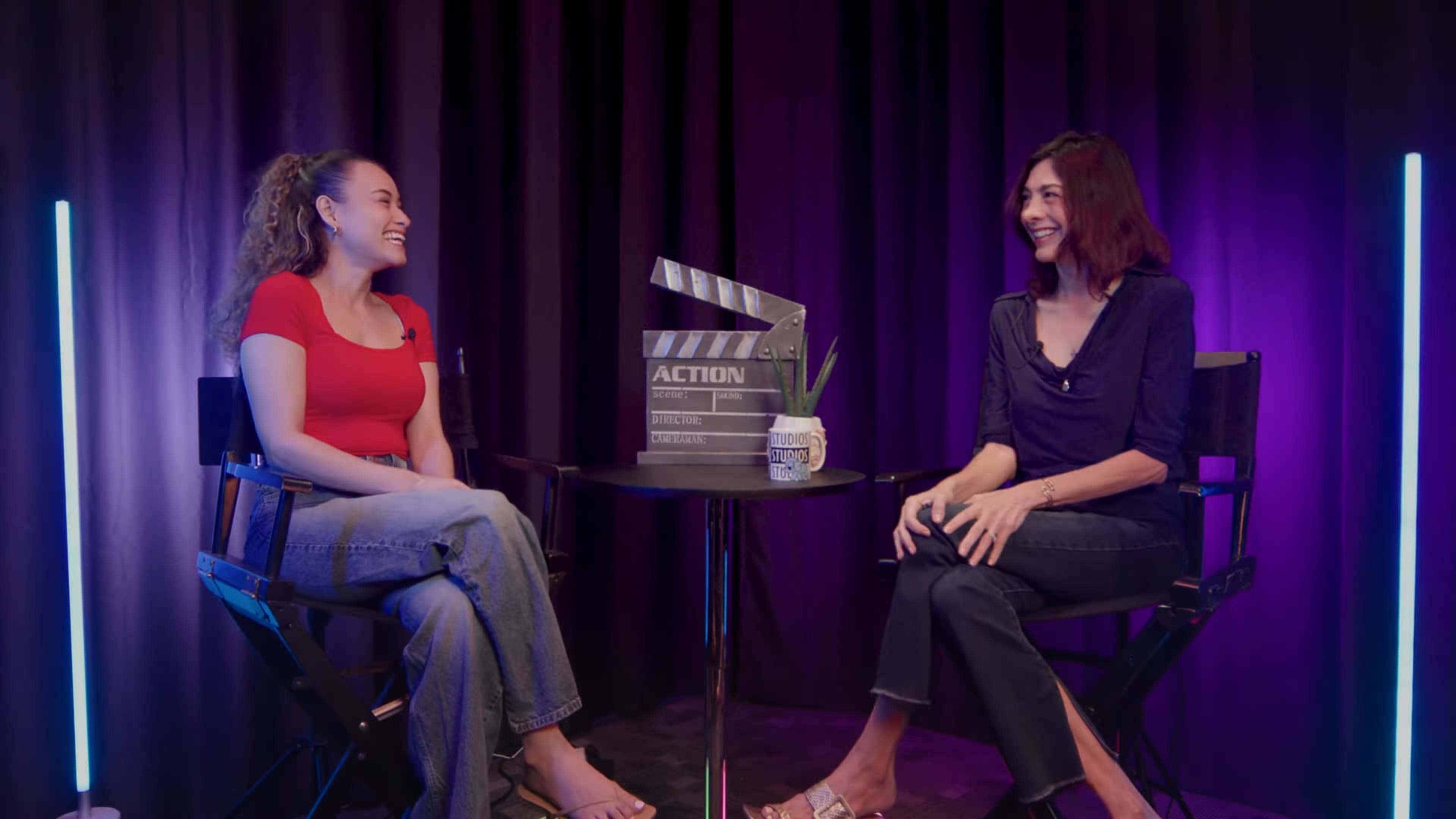 Two women are seated in director's chairs, smiling and conversing, in front of a purple backdrop with a film clapperboard and a plant on the table between them.