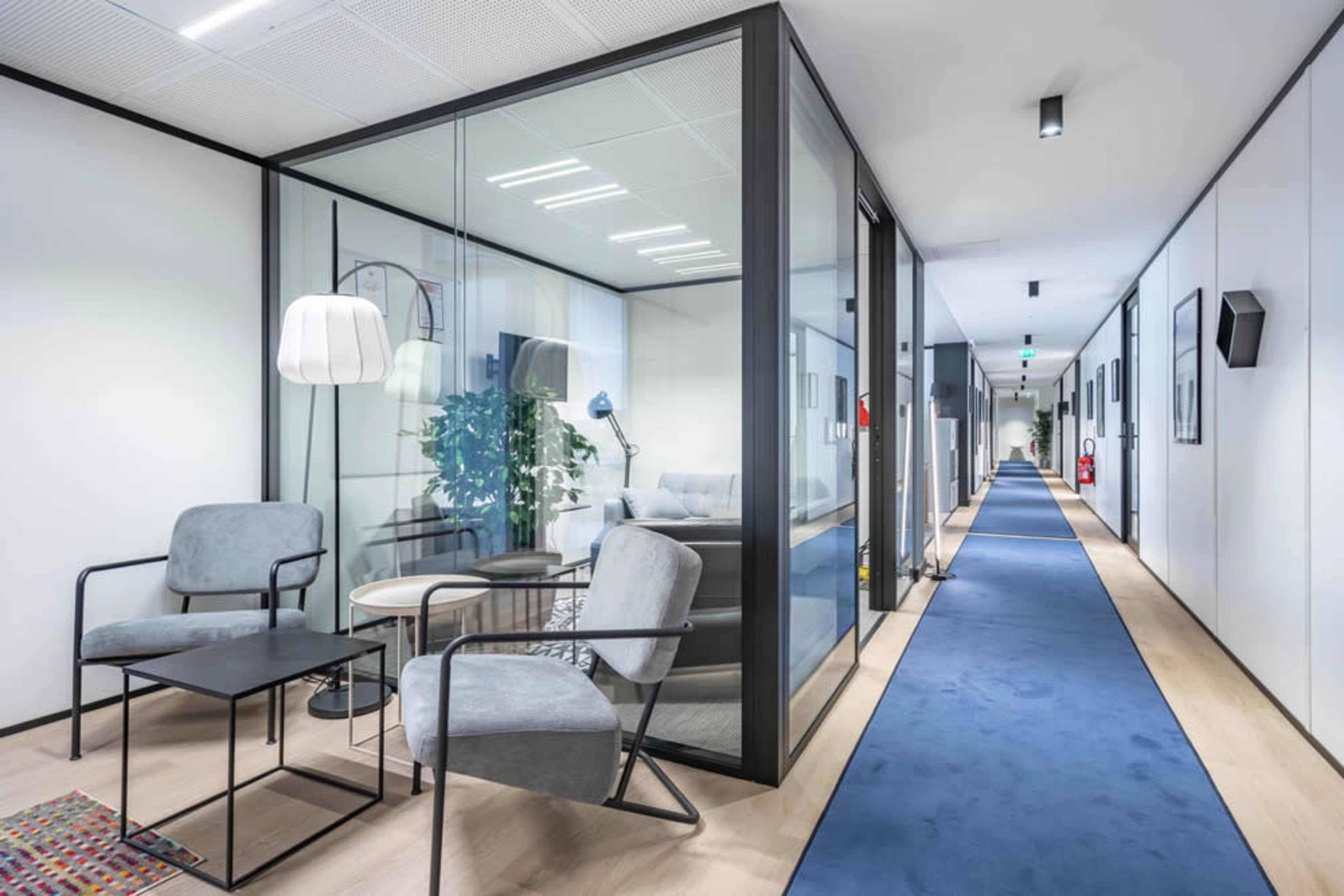 The image shows a modern office hallway featuring glass-walled meeting rooms, blue carpet, and minimalist furniture.