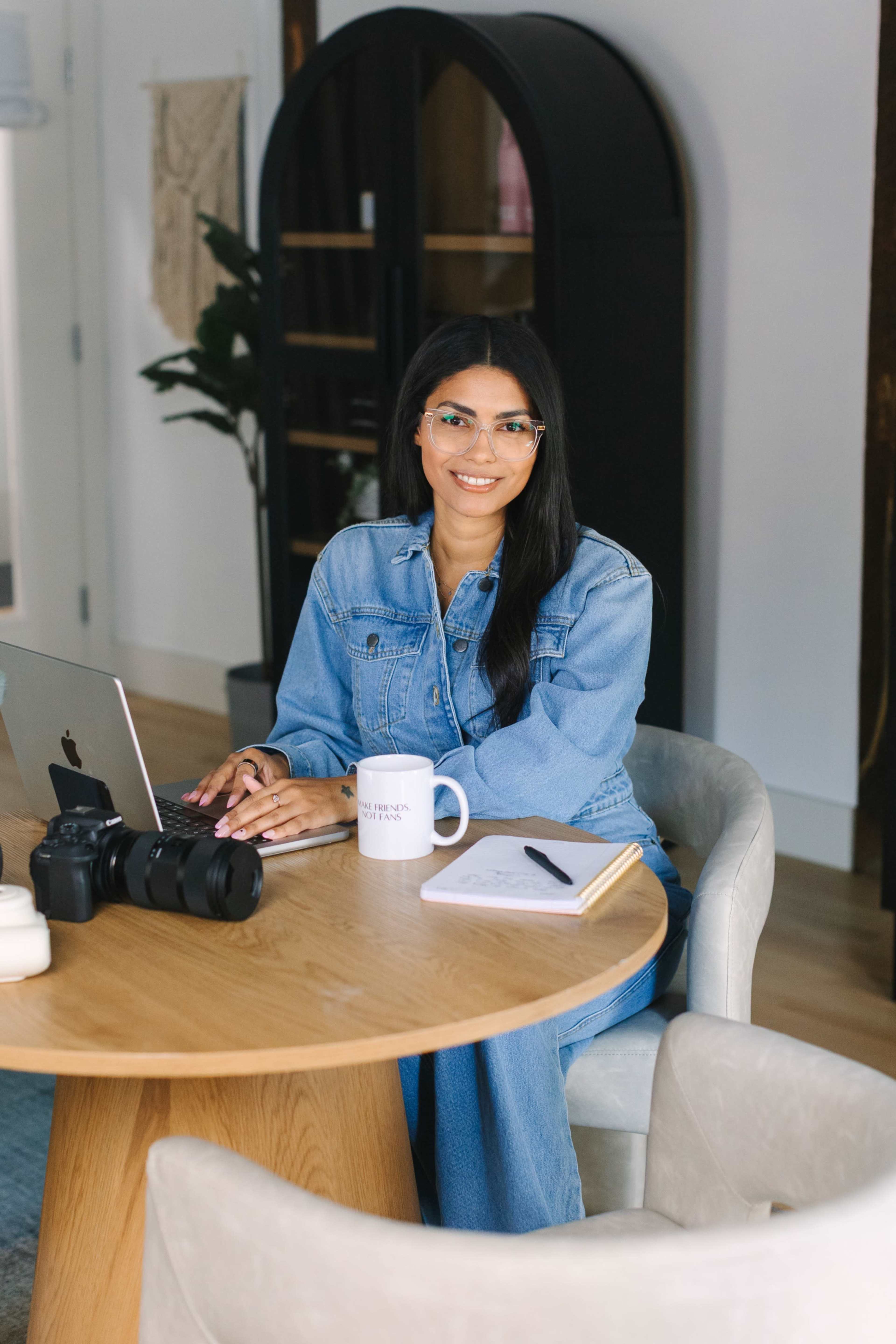 A woman in a denim outfit sits at a wooden table with a laptop, camera, and notebook in a modern office setting.