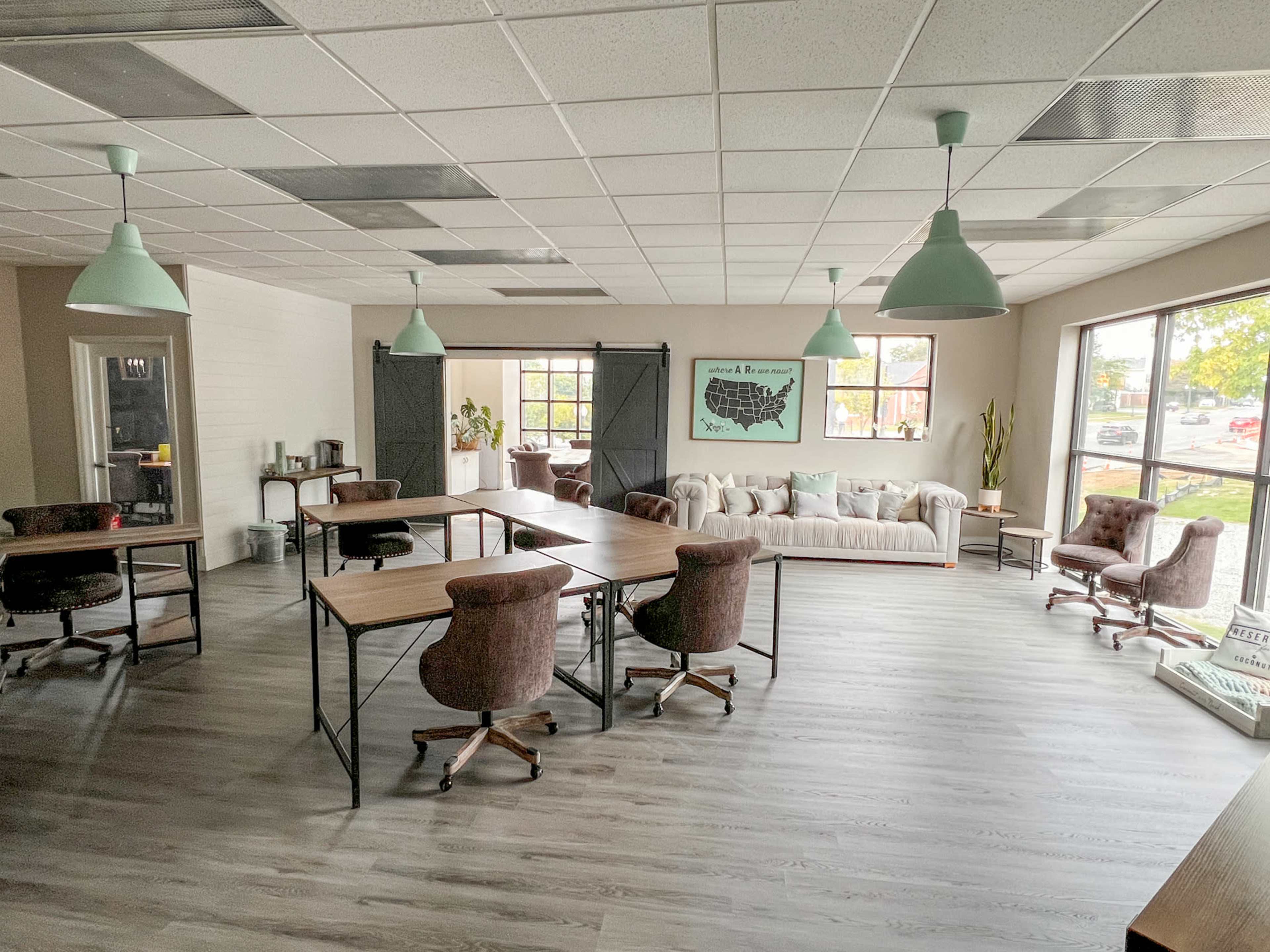 The image shows a modern office space featuring several desks, a comfortable seating area with a sofa, and large windows allowing natural light to enter.