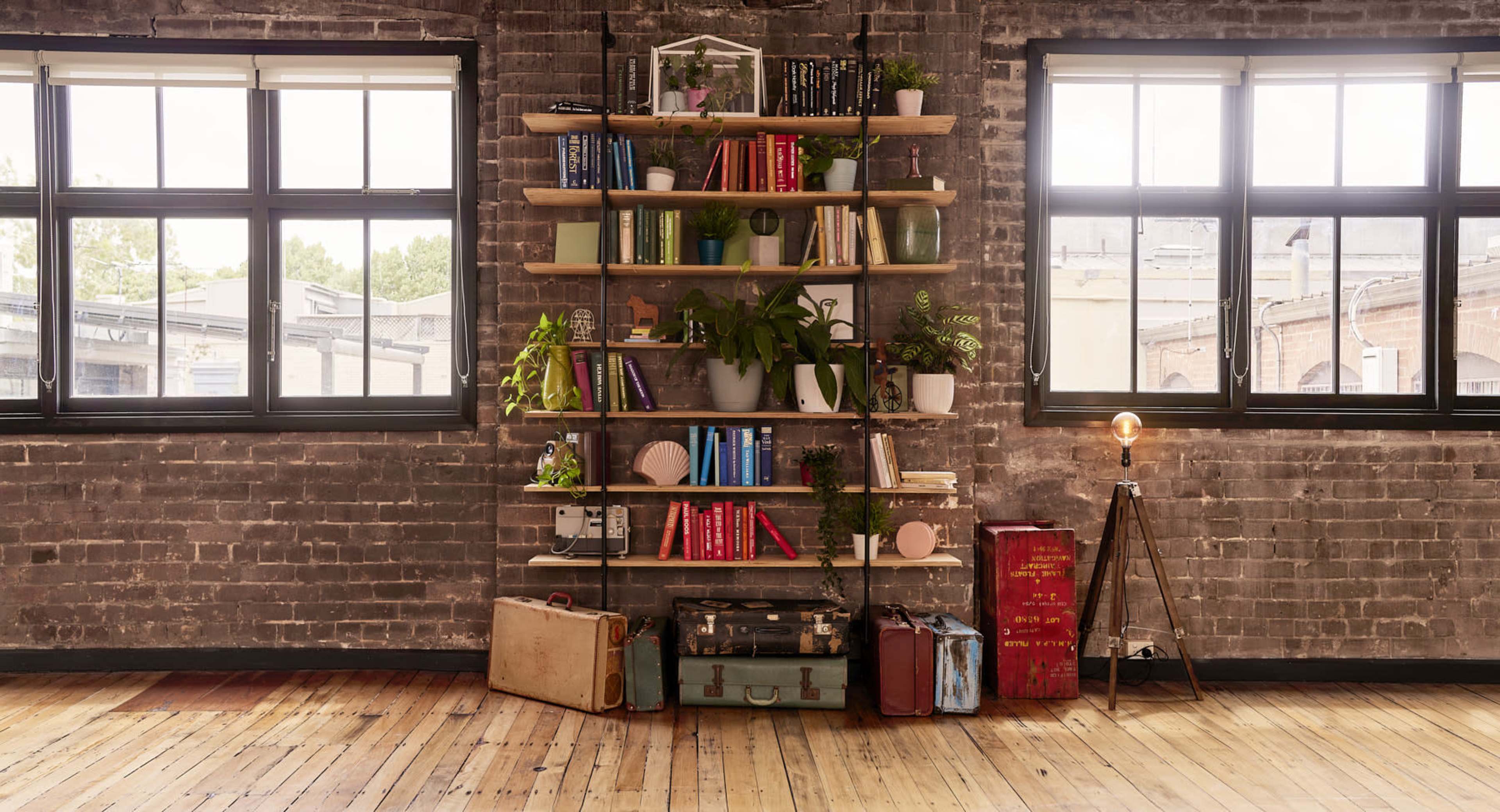 A wooden bookshelf filled with books and plants stands against a brick wall, illuminated by natural light from large windows.