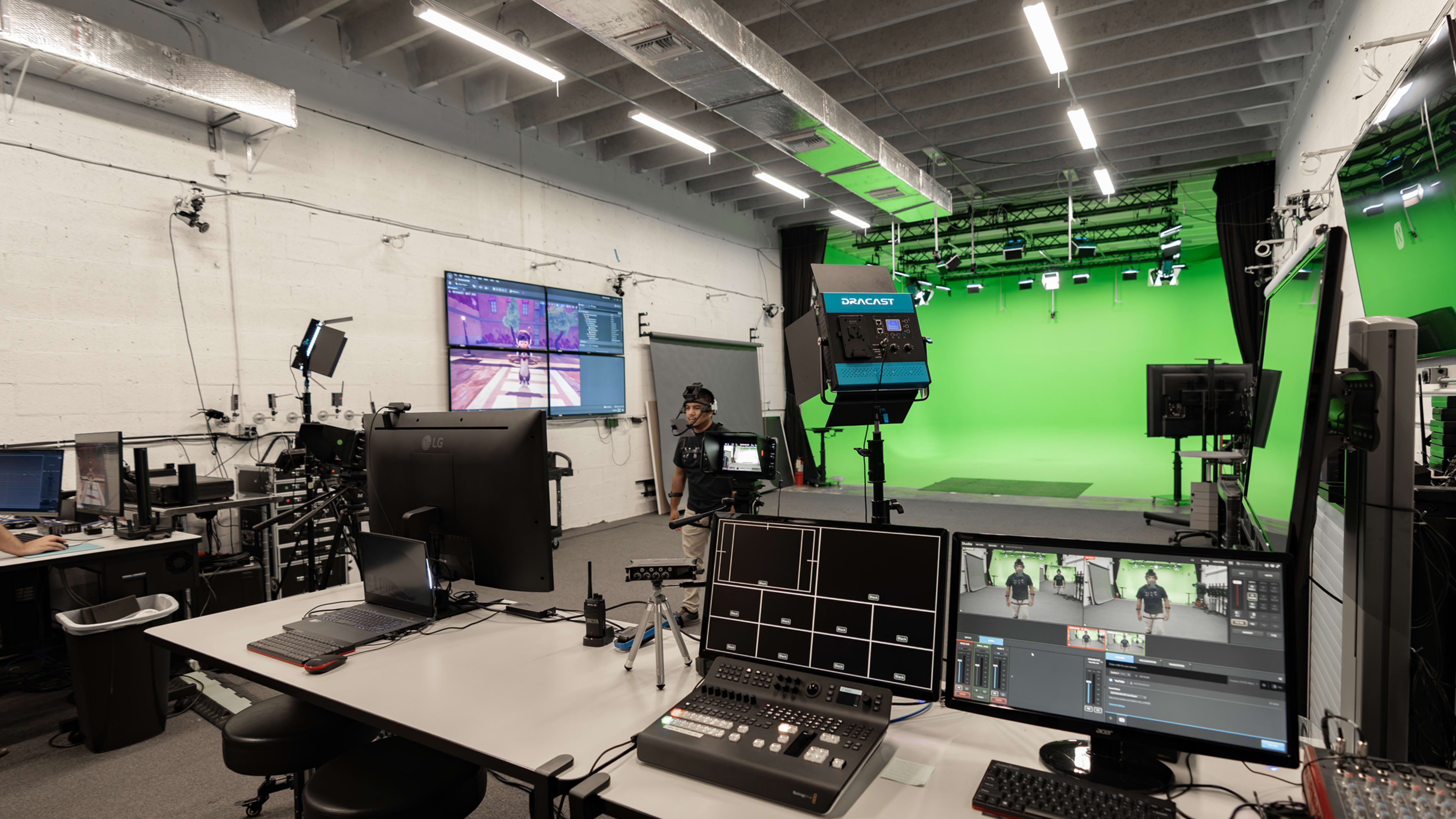 A modern television production studio features multiple computer stations and a green screen backdrop with lighting equipment.