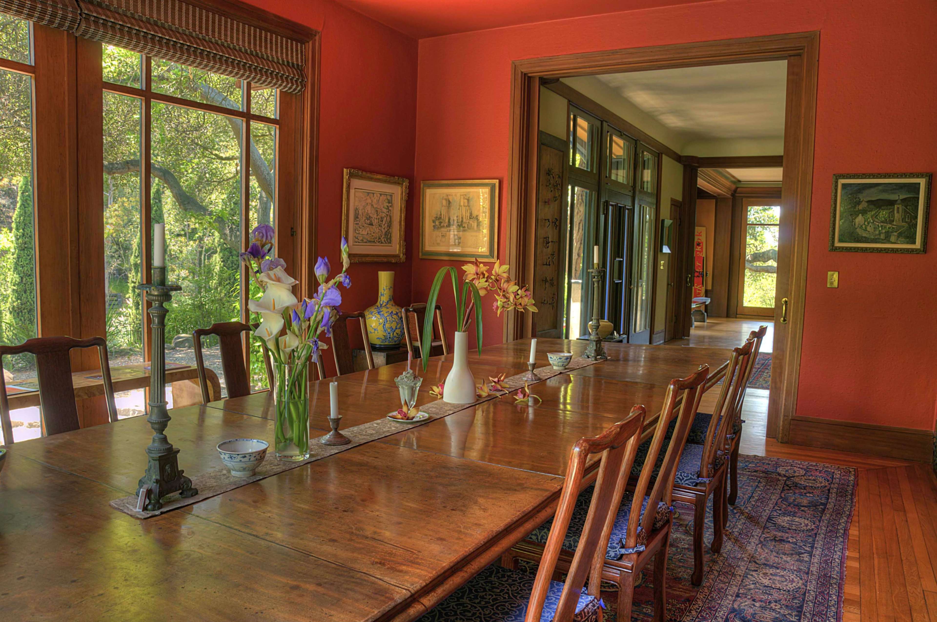 A long wooden dining table is set with vases of flowers in a warmly lit room featuring large windows and colorful walls.