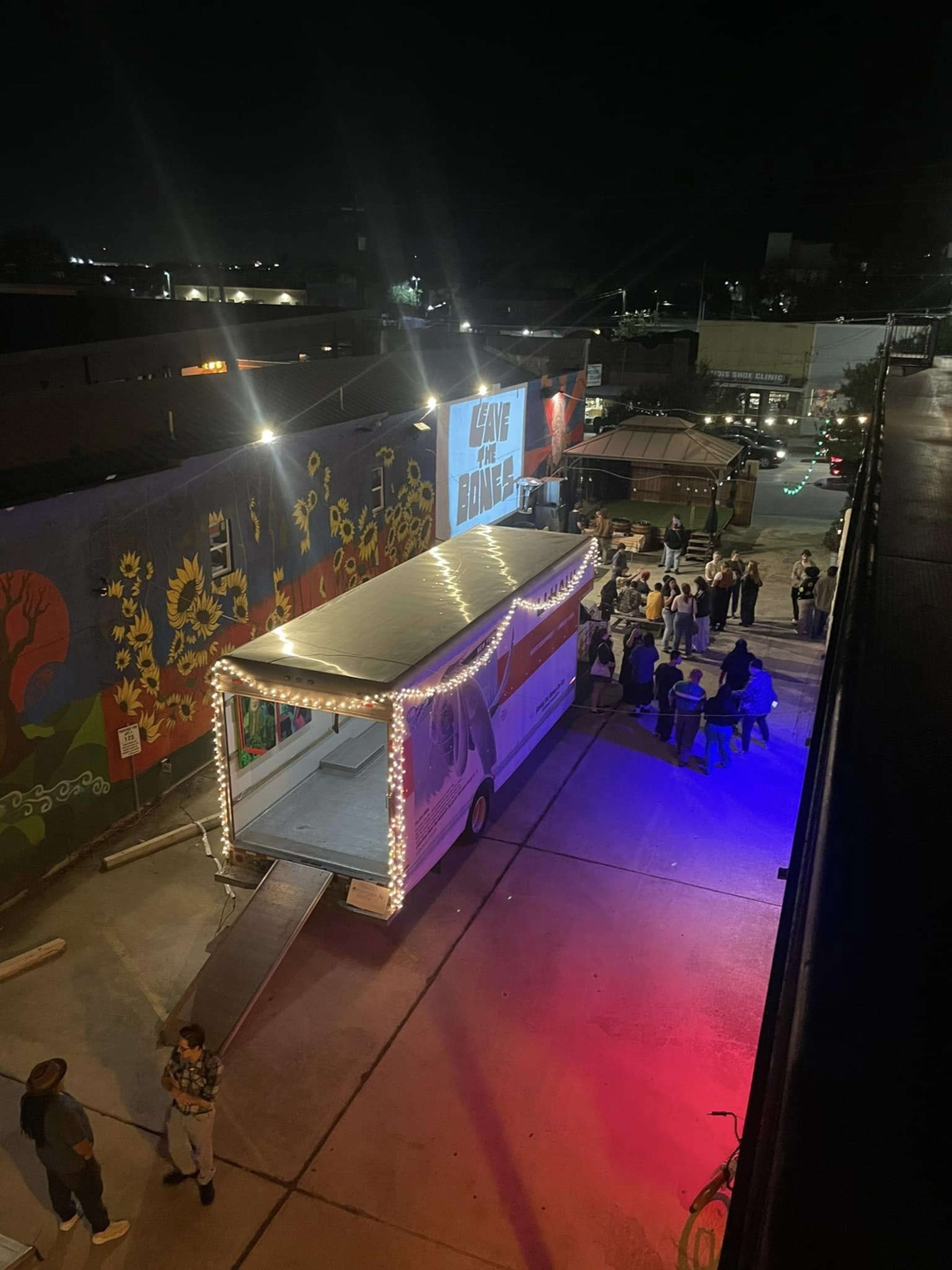 A large food truck is parked in an illuminated alleyway, with people gathered around and a mural on the wall nearby.