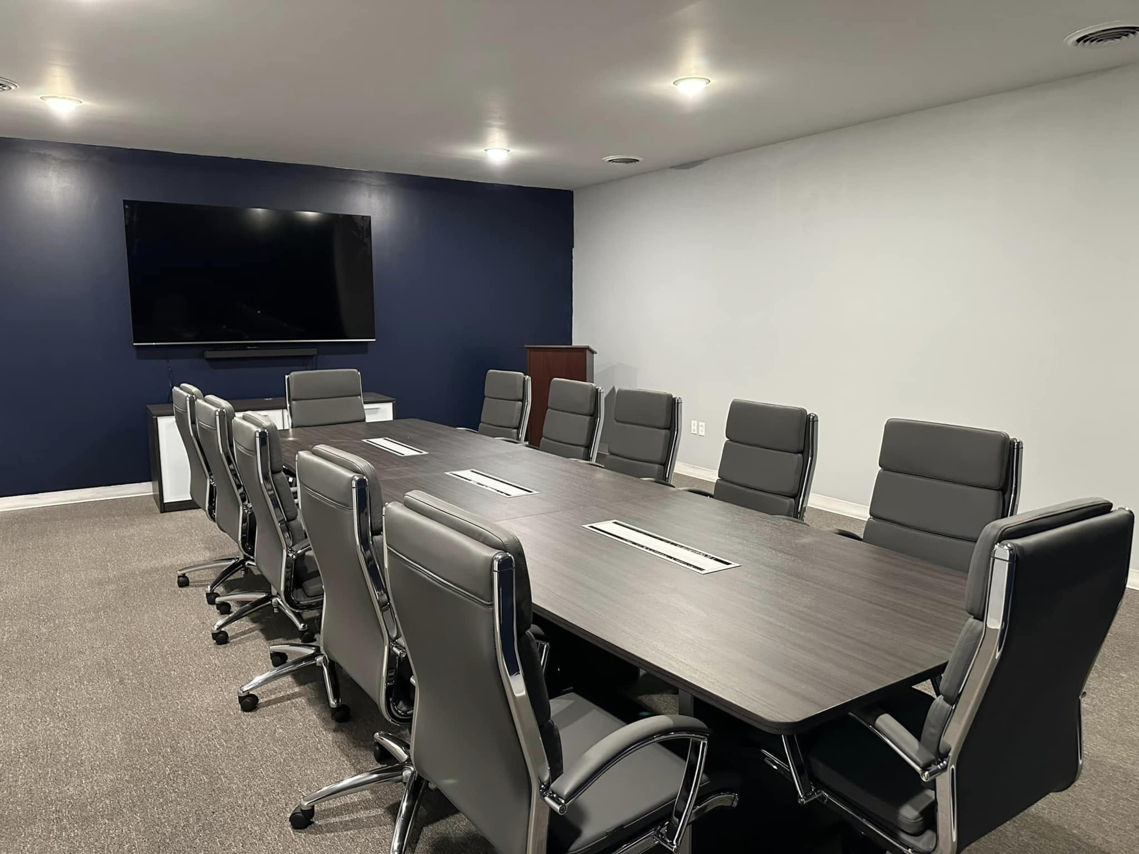 A modern conference room features a long wooden table surrounded by eight ergonomic chairs, with a large screen mounted on the wall.