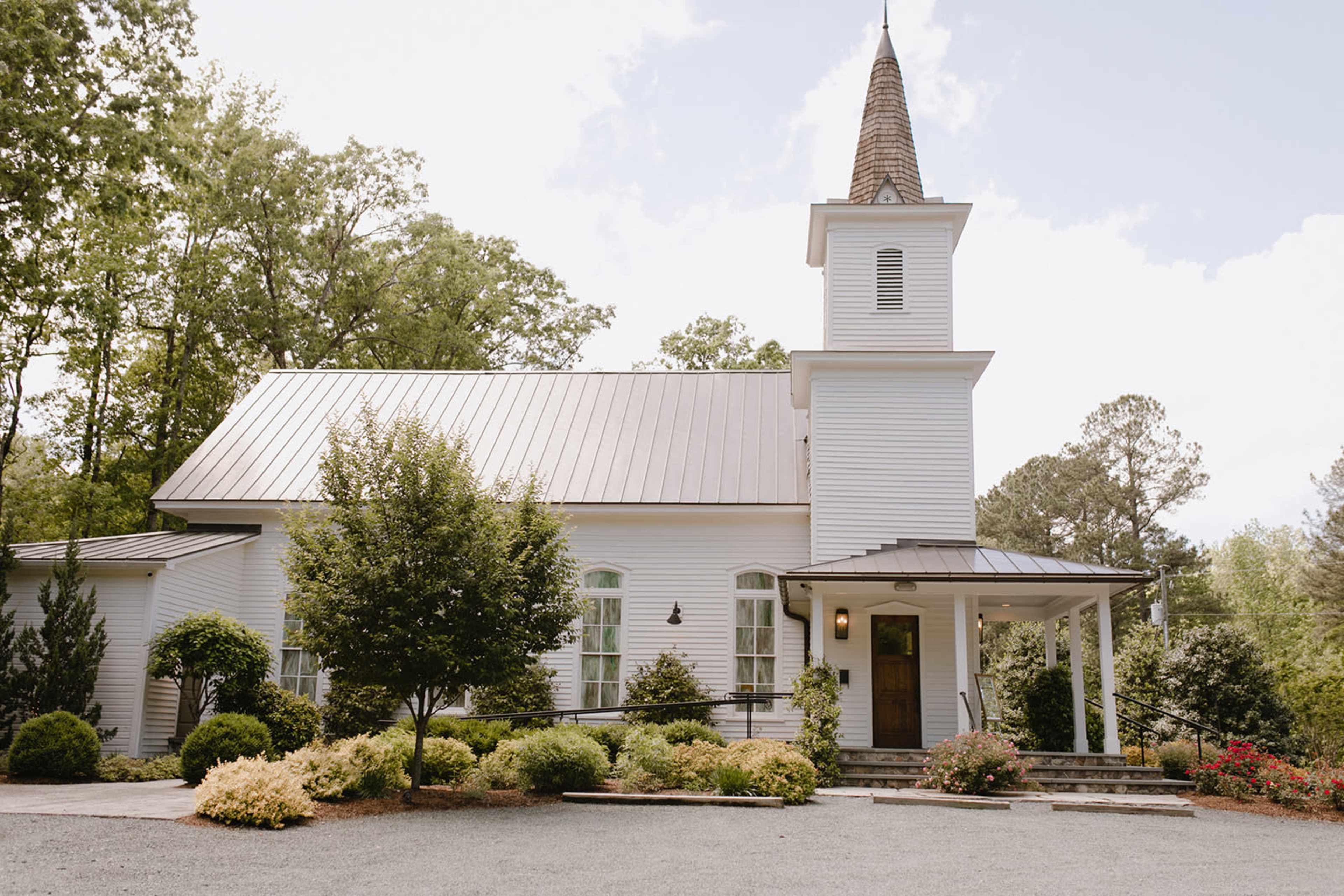 A white church building with a steeple surrounded by green trees and landscaped gardens.