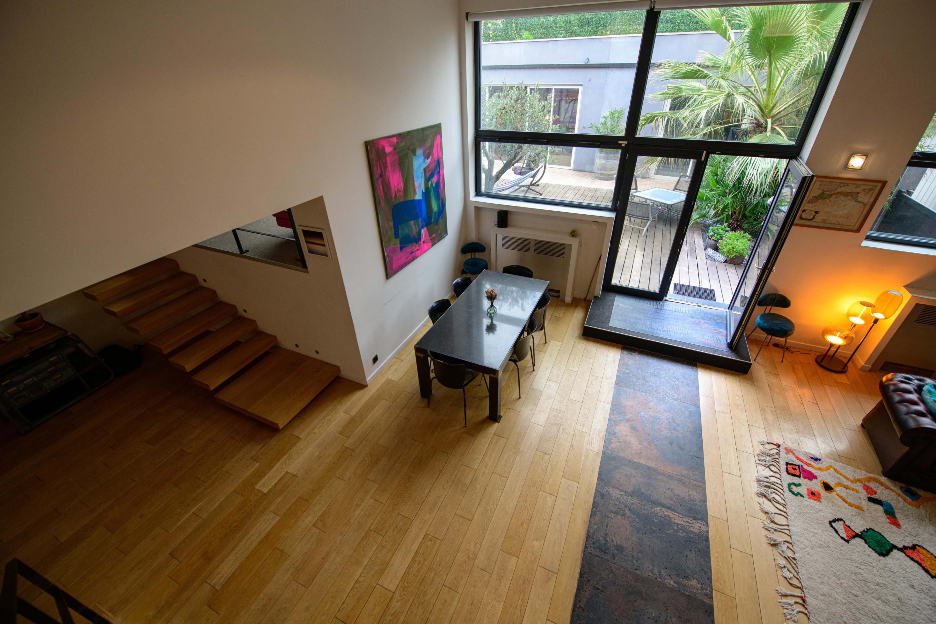 The image shows a modern interior space with a dining area featuring a black table and chairs, large windows allowing natural light, and a staircase leading to an upper level.