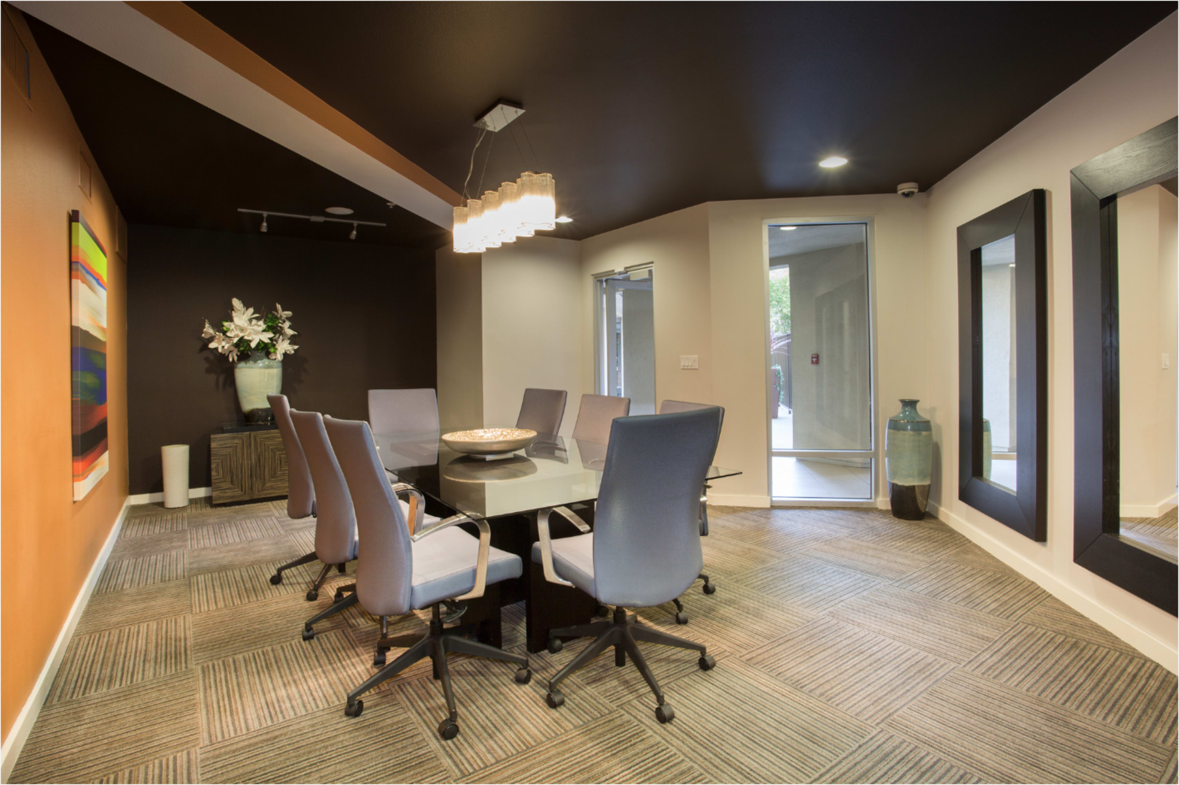 The image shows a modern conference room with a glass table surrounded by six gray chairs, featuring an artwork on the wall and a large mirror opposite the entrance.