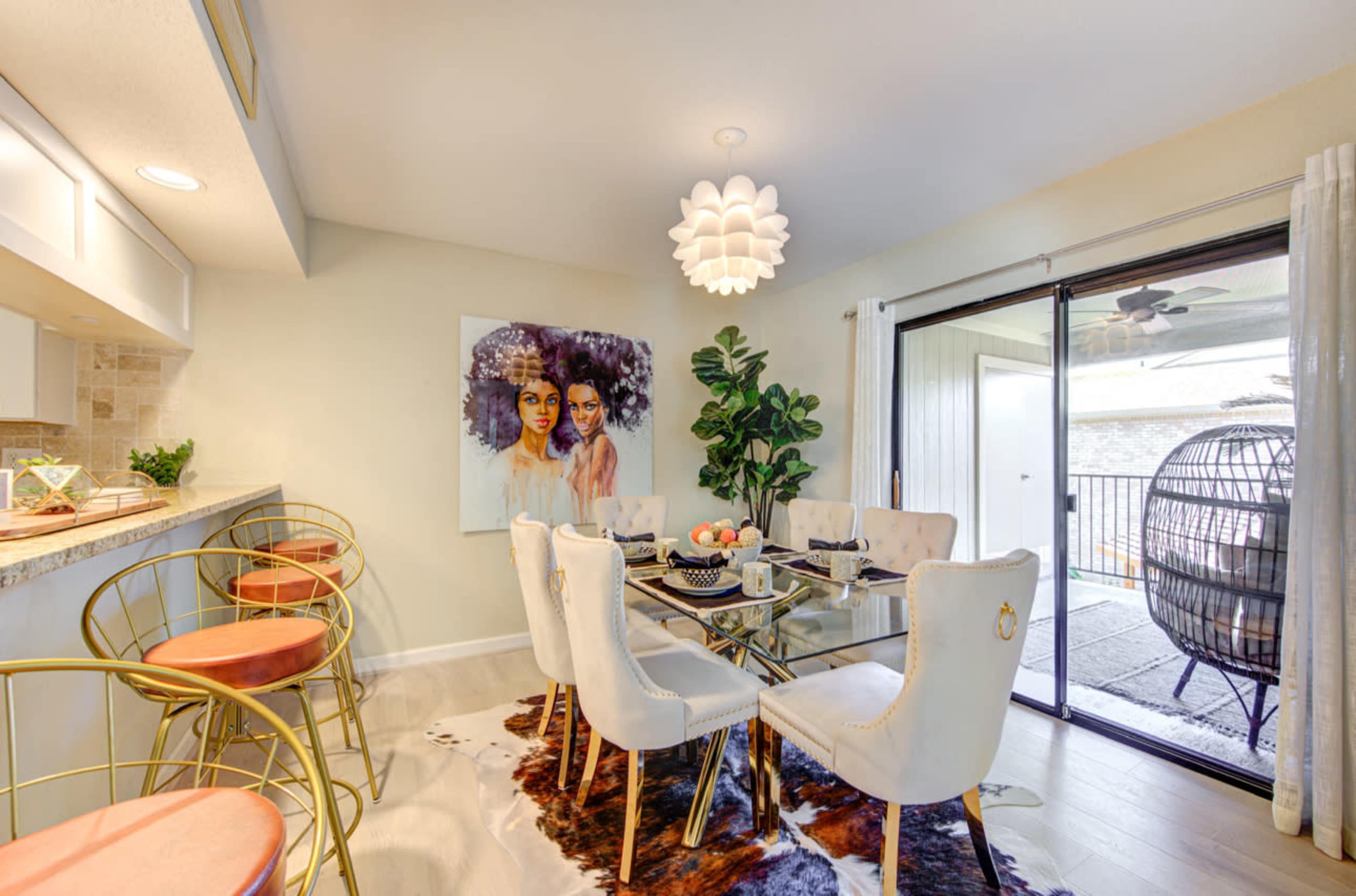 A modern dining area features a glass table surrounded by white chairs, a colorful artwork on the wall, and a sliding glass door leading to a patio.