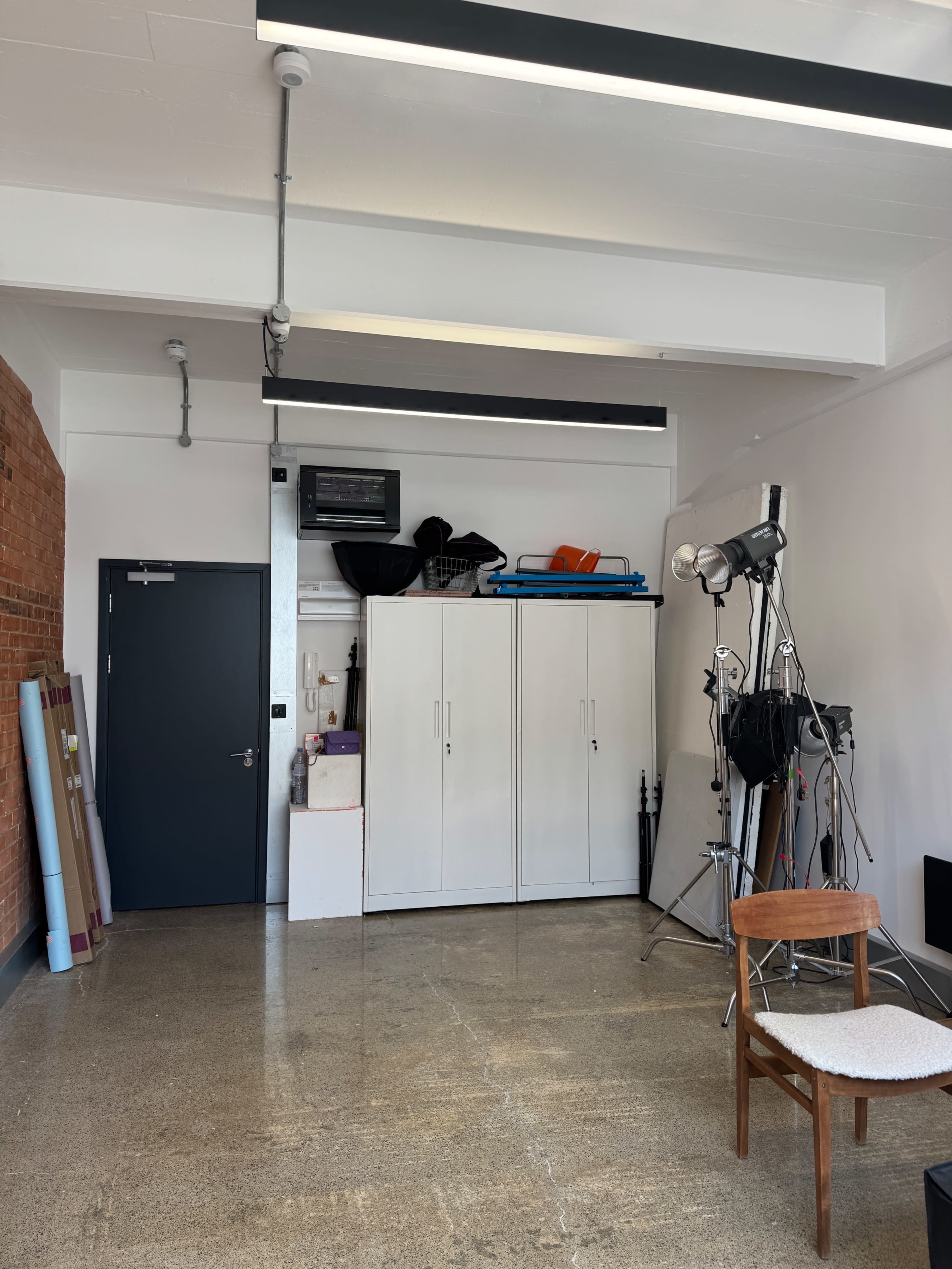 The image shows a spacious studio with exposed brick walls, storage cabinets, various equipment, and a chair in the foreground.