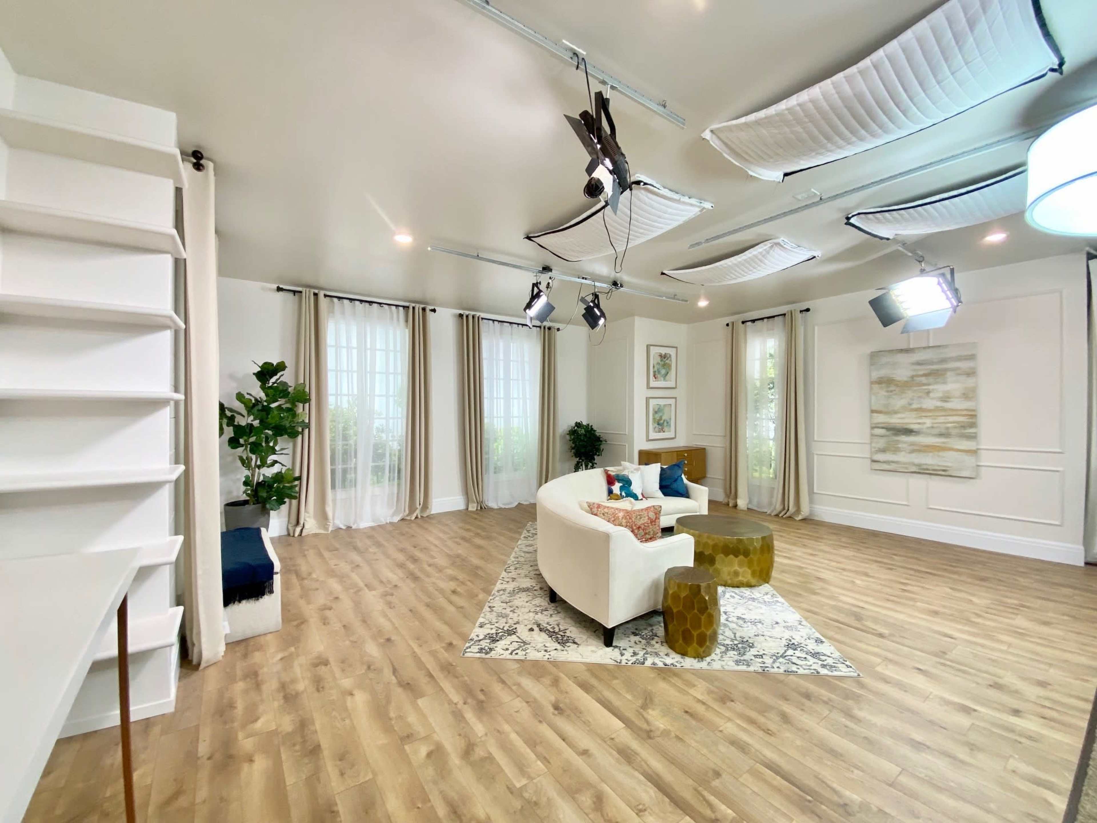 A modern, well-lit studio interior featuring a white sectional sofa, a patterned area rug, and decorative plants against light-colored walls.