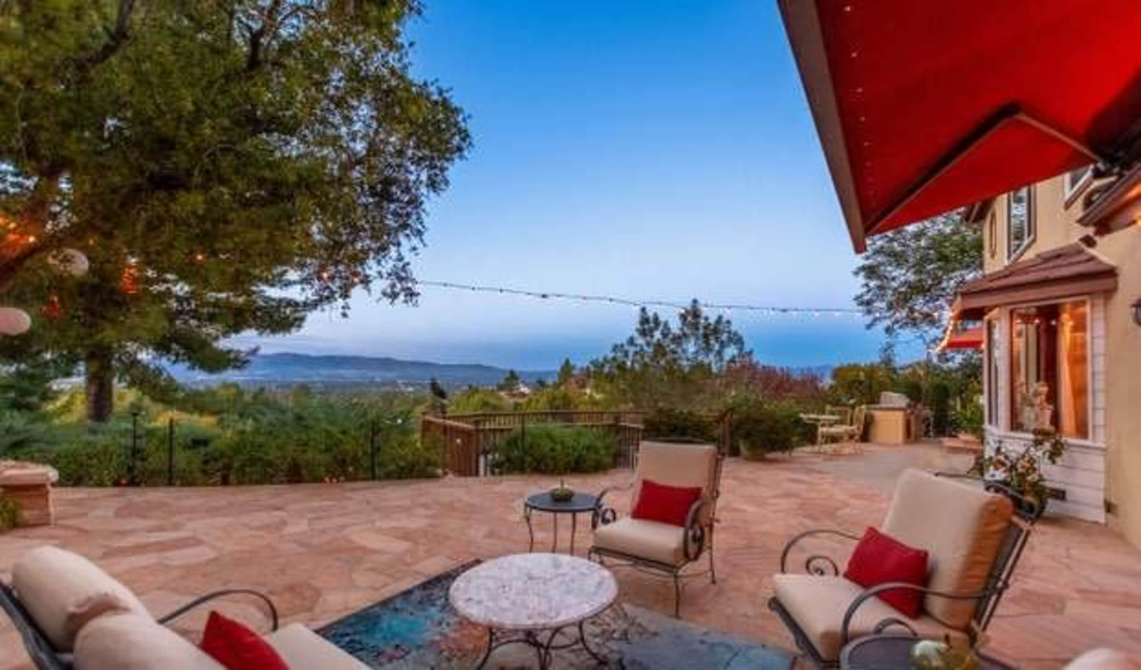 A patio area features two lounge chairs with cushions, a round table, and a distant view of rolling hills under a clear sky.