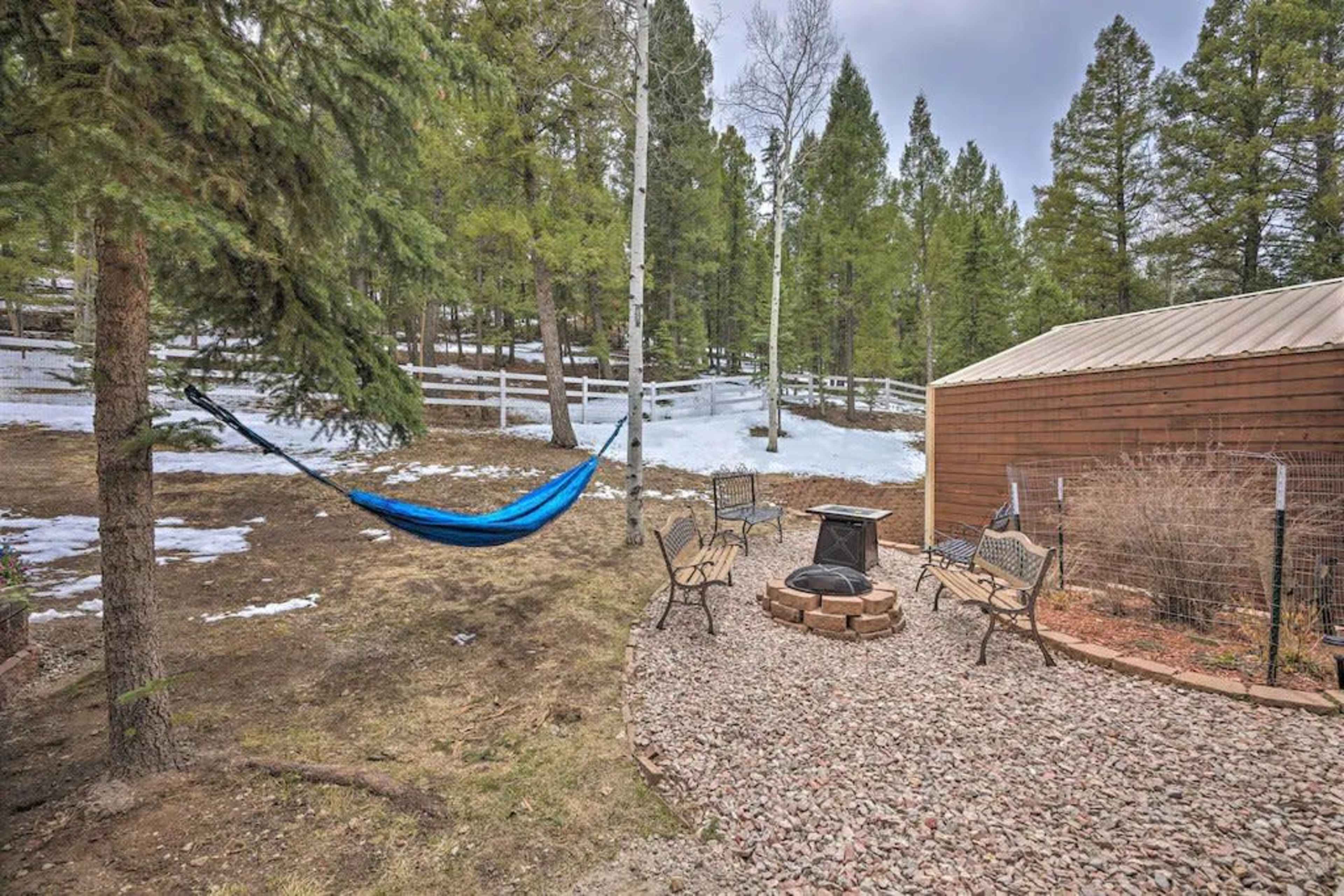 A blue hammock is suspended between two trees near a stone fire pit surrounded by seating in a snow-covered wooded area.