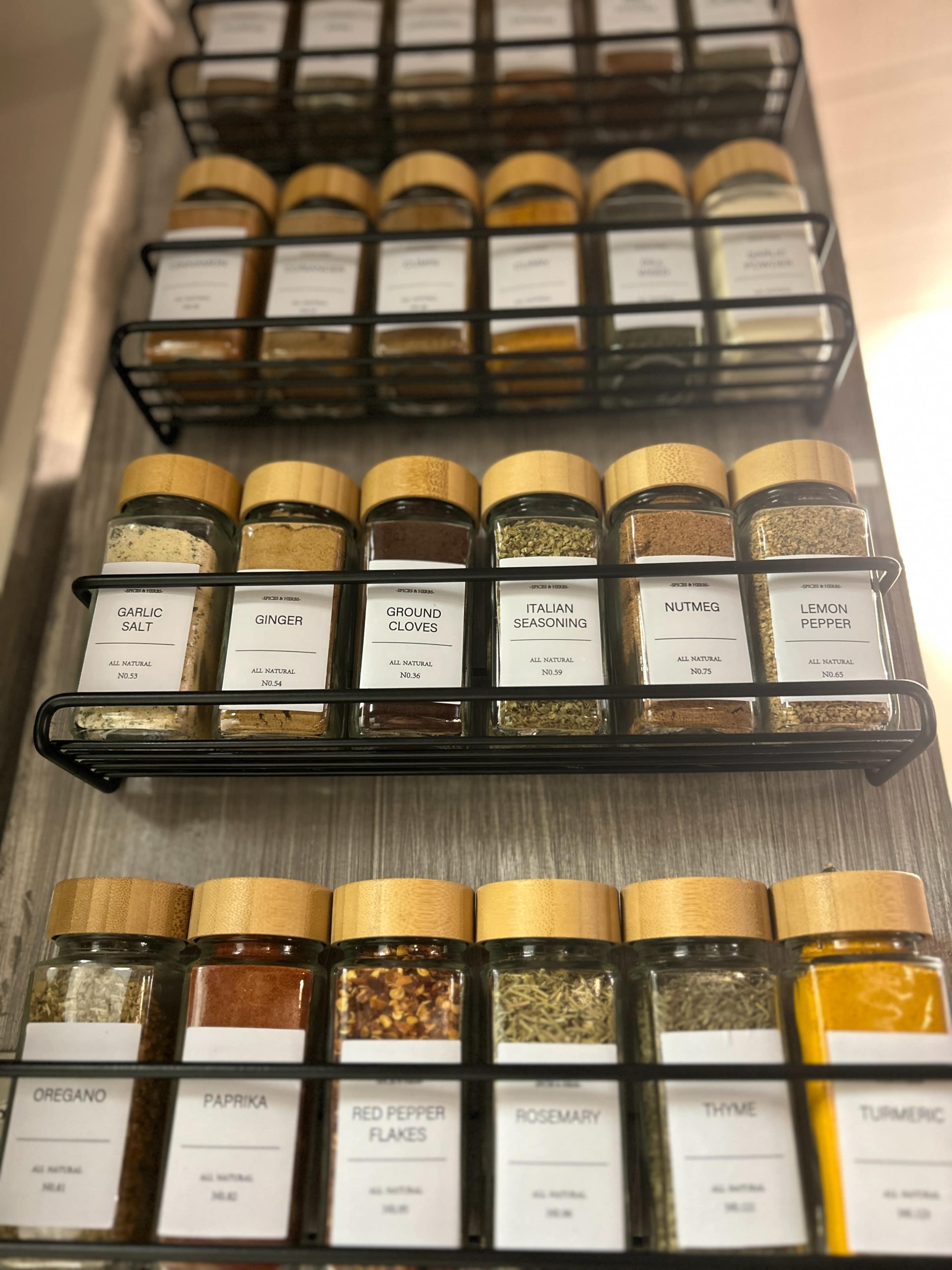 A rack holds various labeled spice jars with wooden lids arranged in rows.