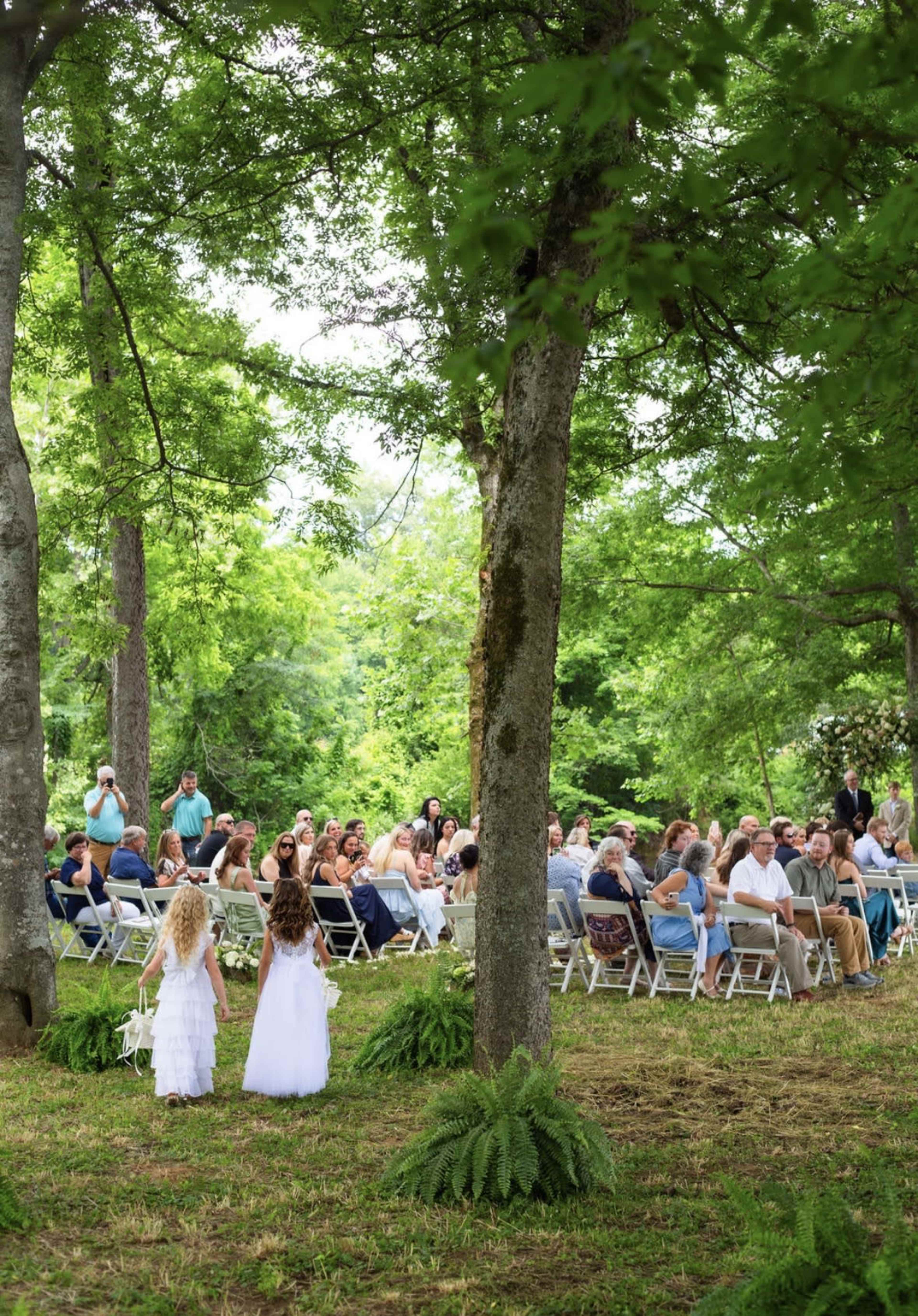 A wedding ceremony is taking place outdoors under trees, with guests seated in white chairs facing two young girls in white dresses.
