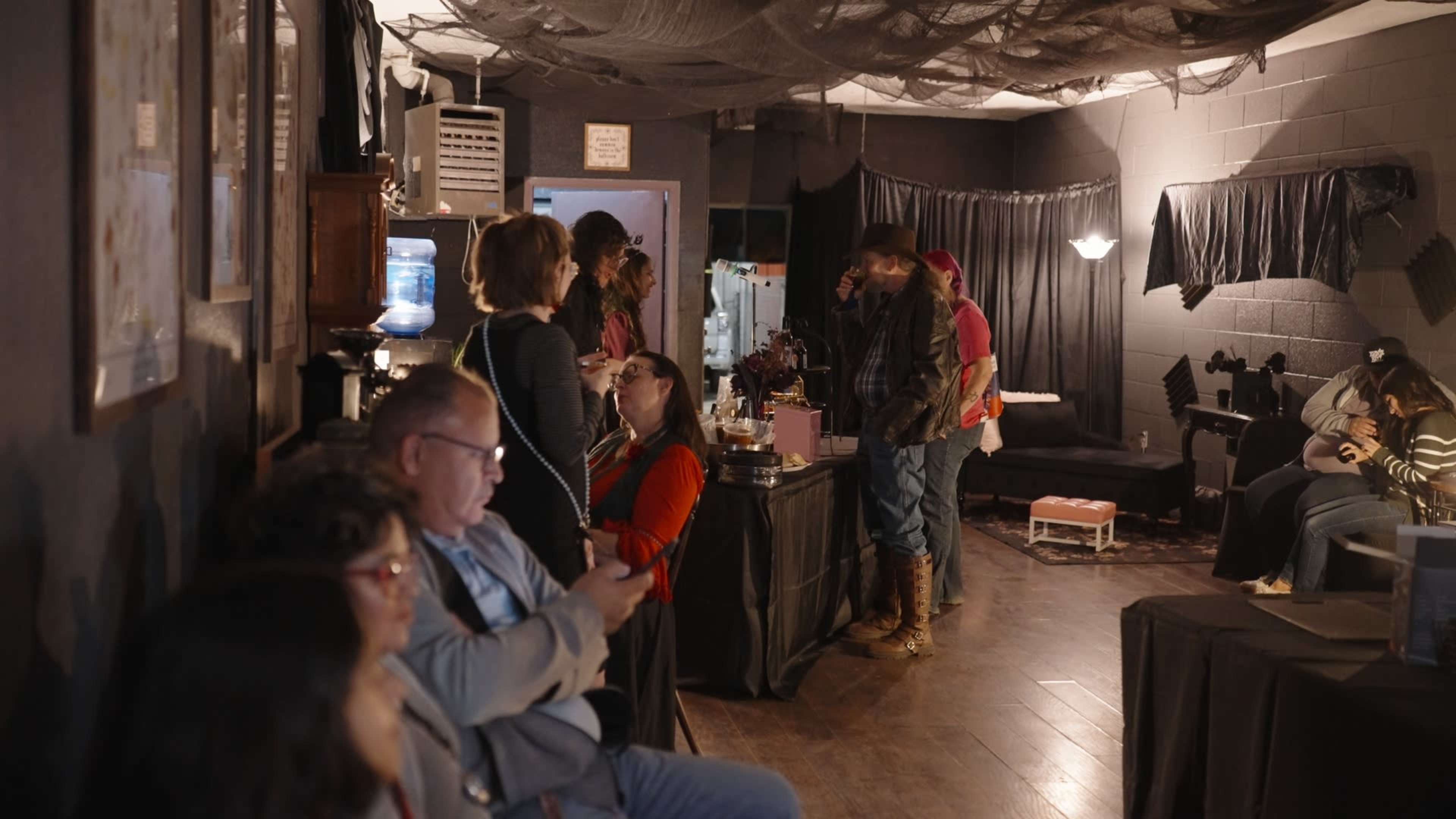 A group of people socialize in a dimly lit room decorated with black drapes and eerie decorations, while some sit on benches and others stand nearby.