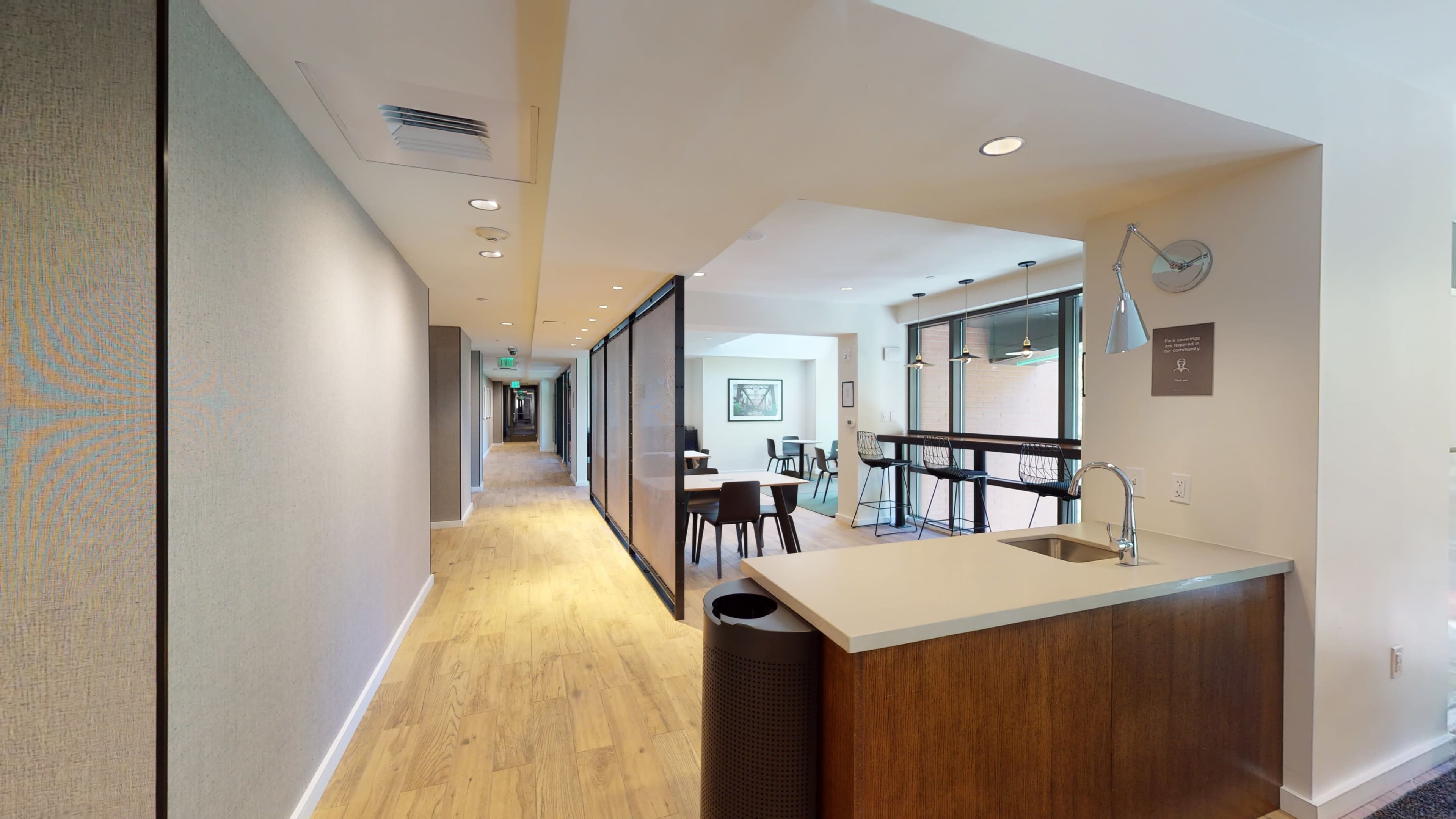 A modern hallway with wooden flooring, leading to various rooms, featuring glass partitions and a small kitchenette on one side.