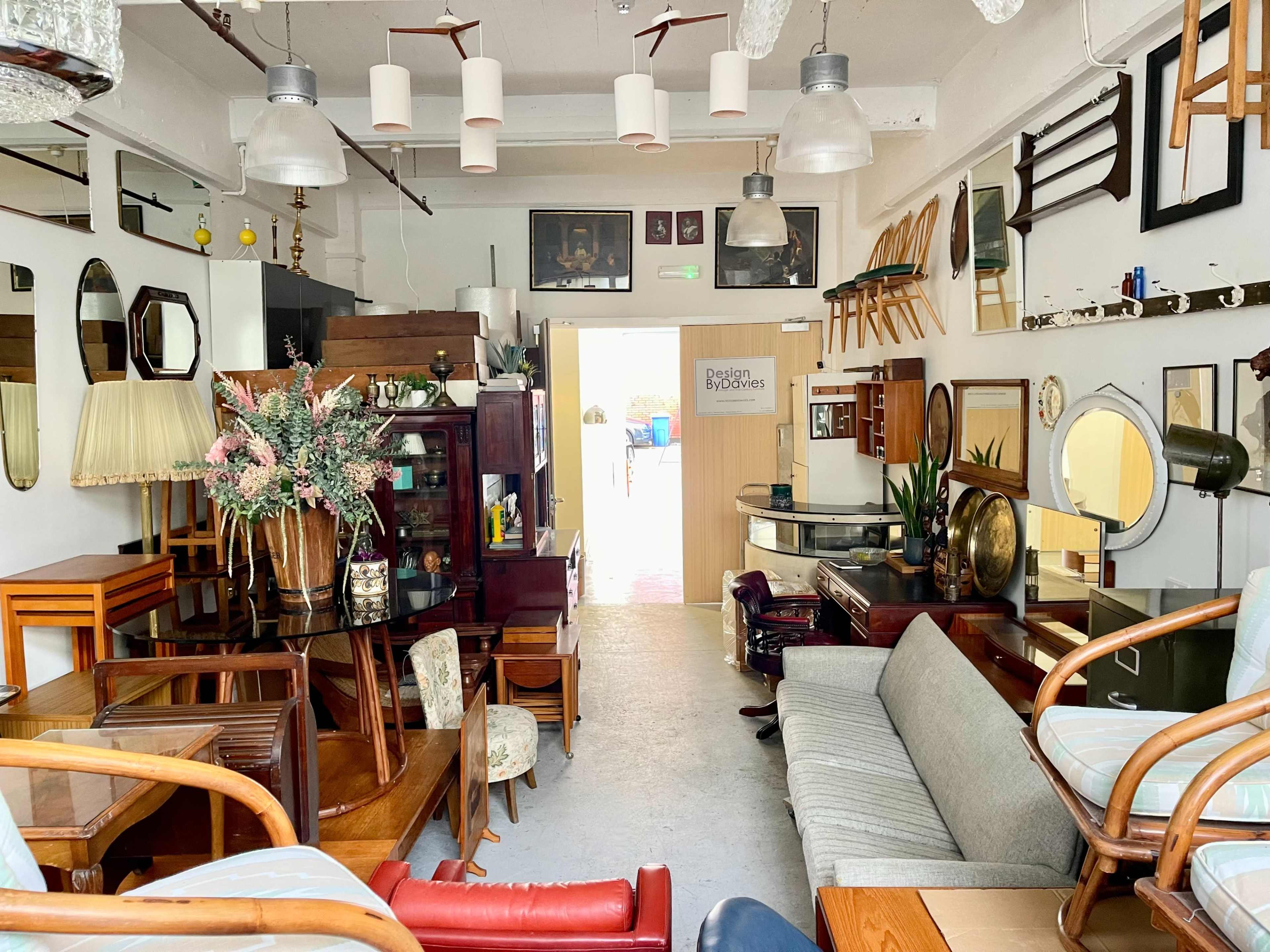 The image shows a vintage furniture store filled with various wooden tables, chairs, mirrors, and decorative items arranged throughout the space.