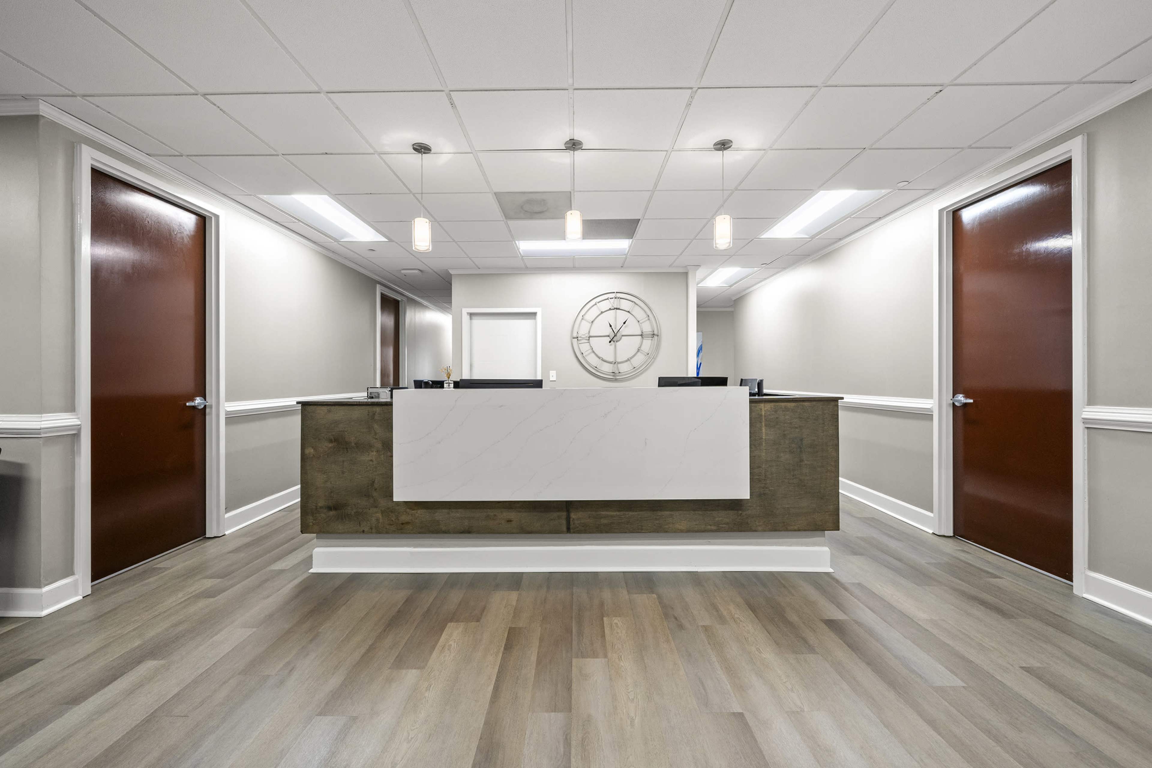The image shows a modern reception area with a wooden front desk, two closed red doors on either side, and a wall clock above the desk.