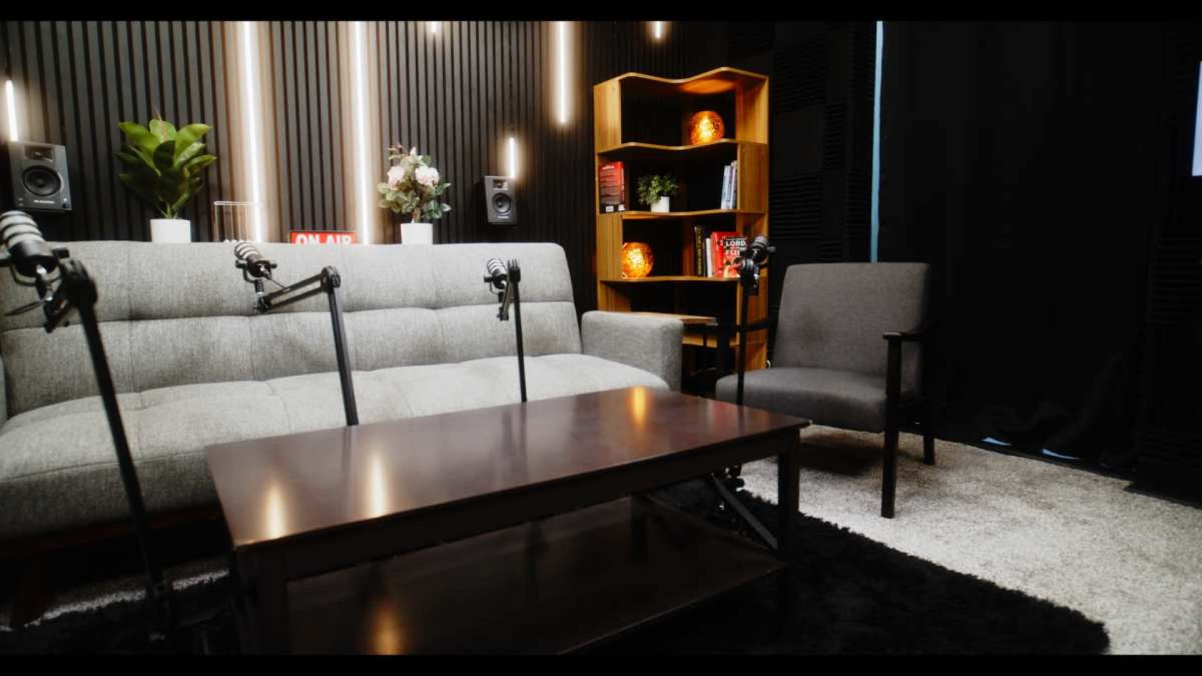 The image shows a modern podcast studio featuring a gray sofa, a wooden coffee table, two microphones on stands, and decorative lighting on a black wall.