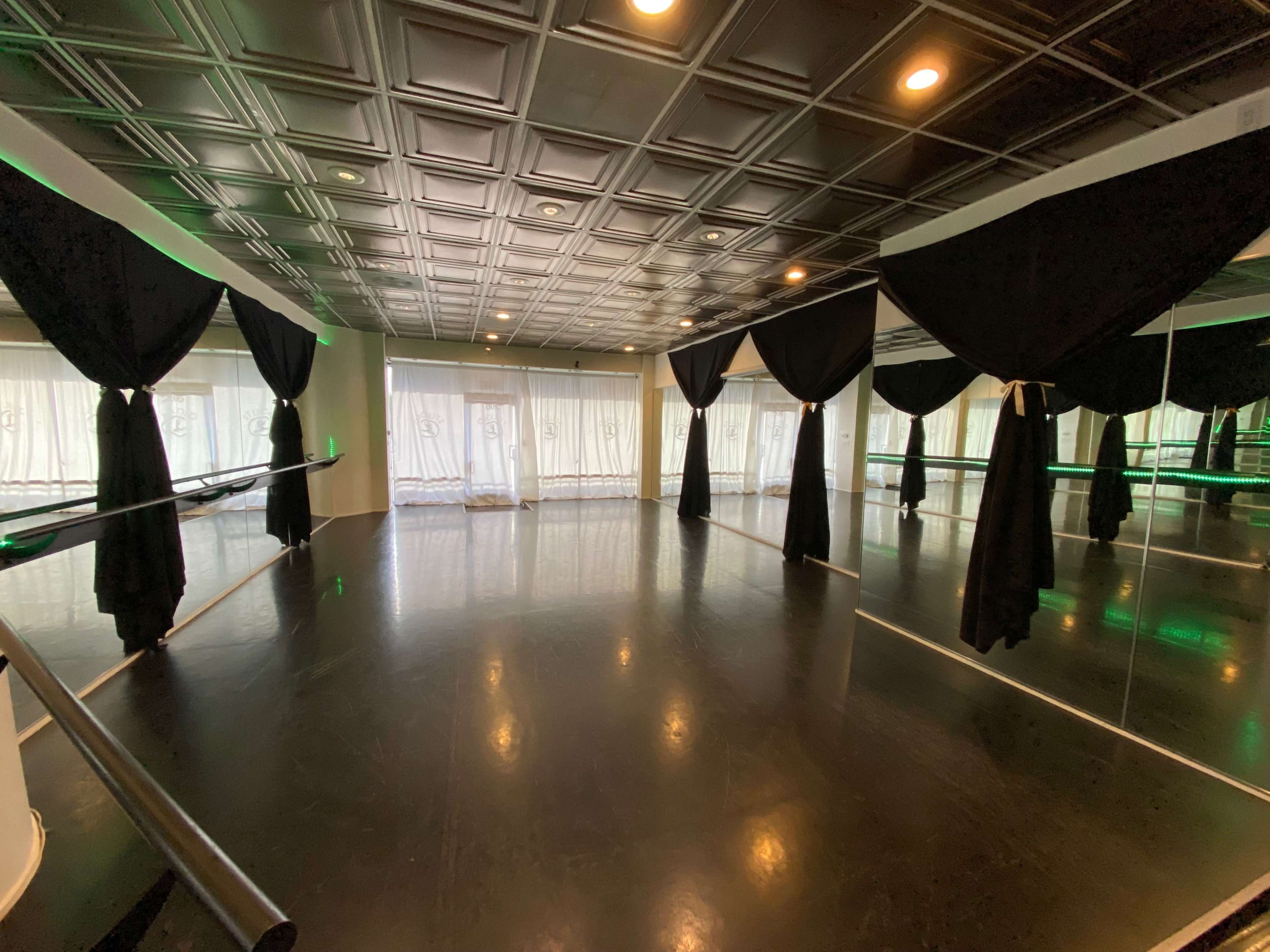 A dance studio with a polished black floor, large windows draped with sheer white curtains, and black curtains hanging from the ceiling.