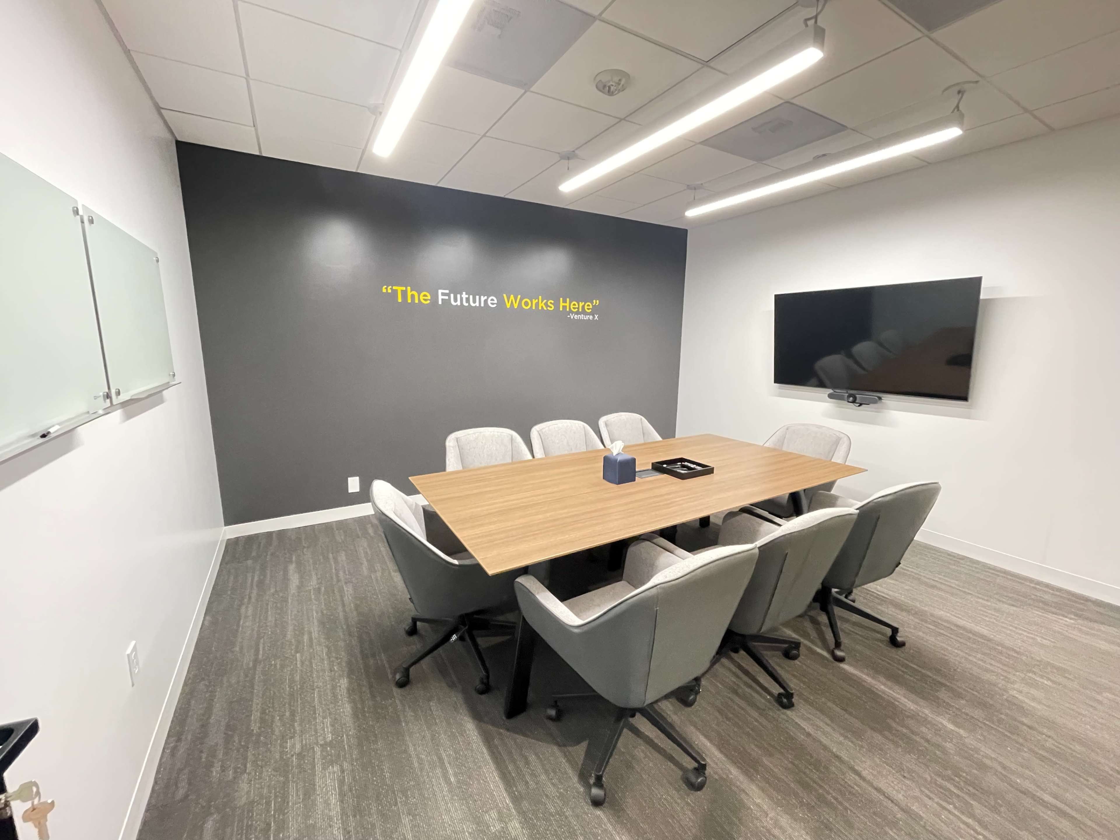 A modern conference room features a large wooden table surrounded by six upholstered chairs, with a wall-mounted screen and a motivational quote displayed above.