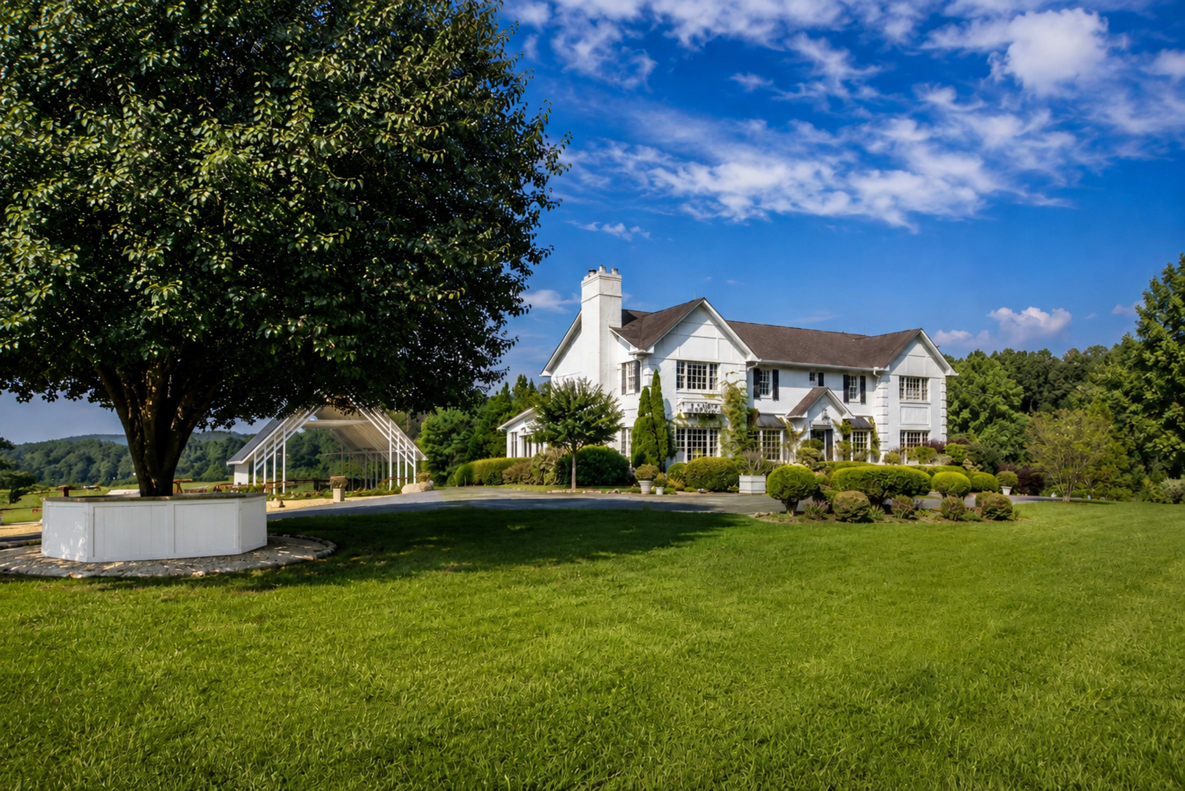 A large white two-story house with a grassy front yard and a tree stands under a blue sky with scattered clouds.