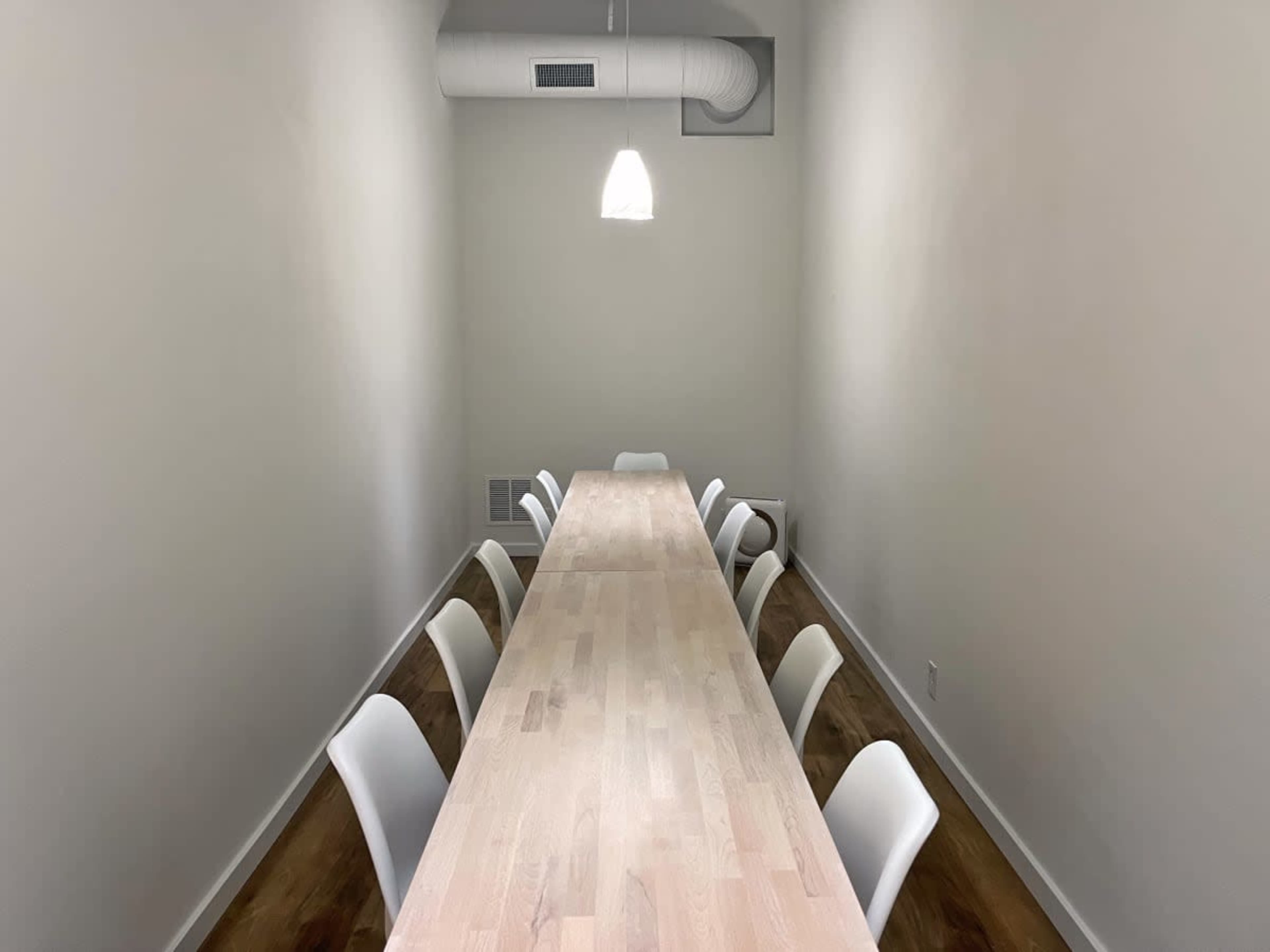 A long wooden table with white chairs is positioned in a well-lit, minimalist meeting room.
