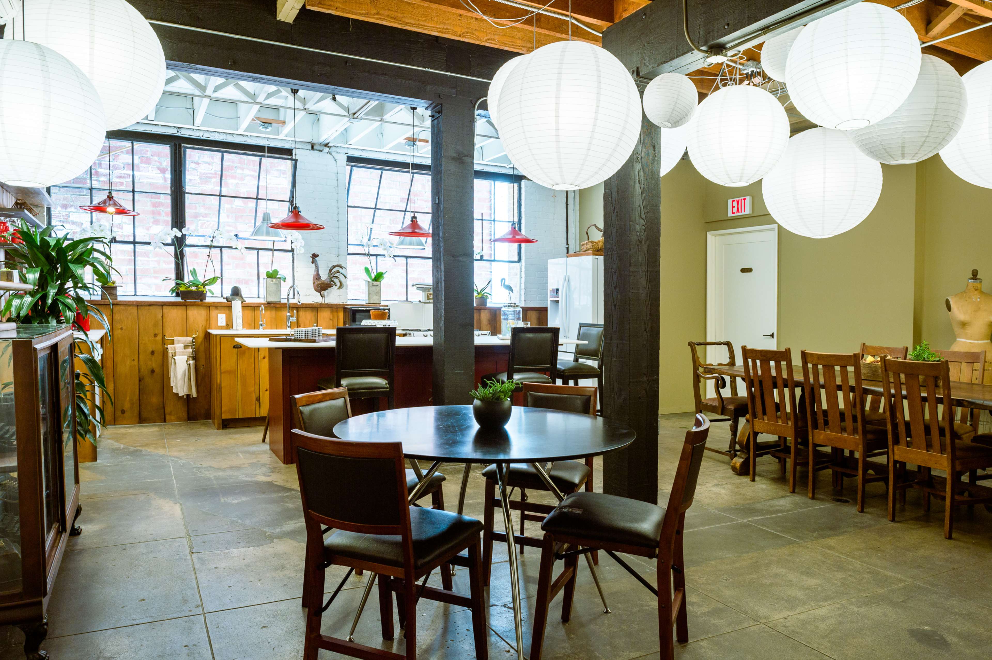 A spacious dining area with a round table and chairs, surrounded by a kitchen, pendant lights, and potted plants in a well-lit room.