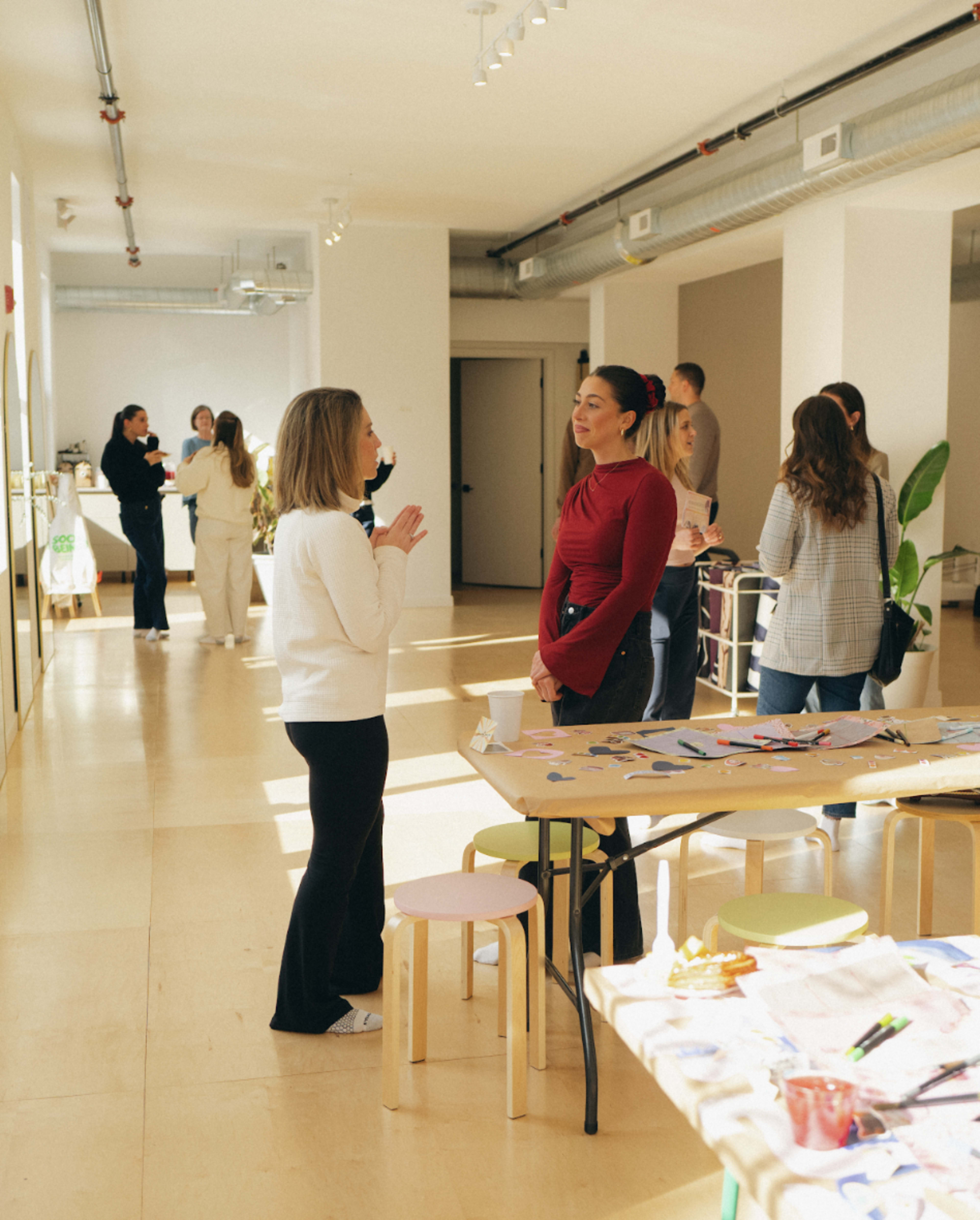 Two women are engaged in conversation in a well-lit, modern space with several people mingling in the background and tables covered with art supplies.