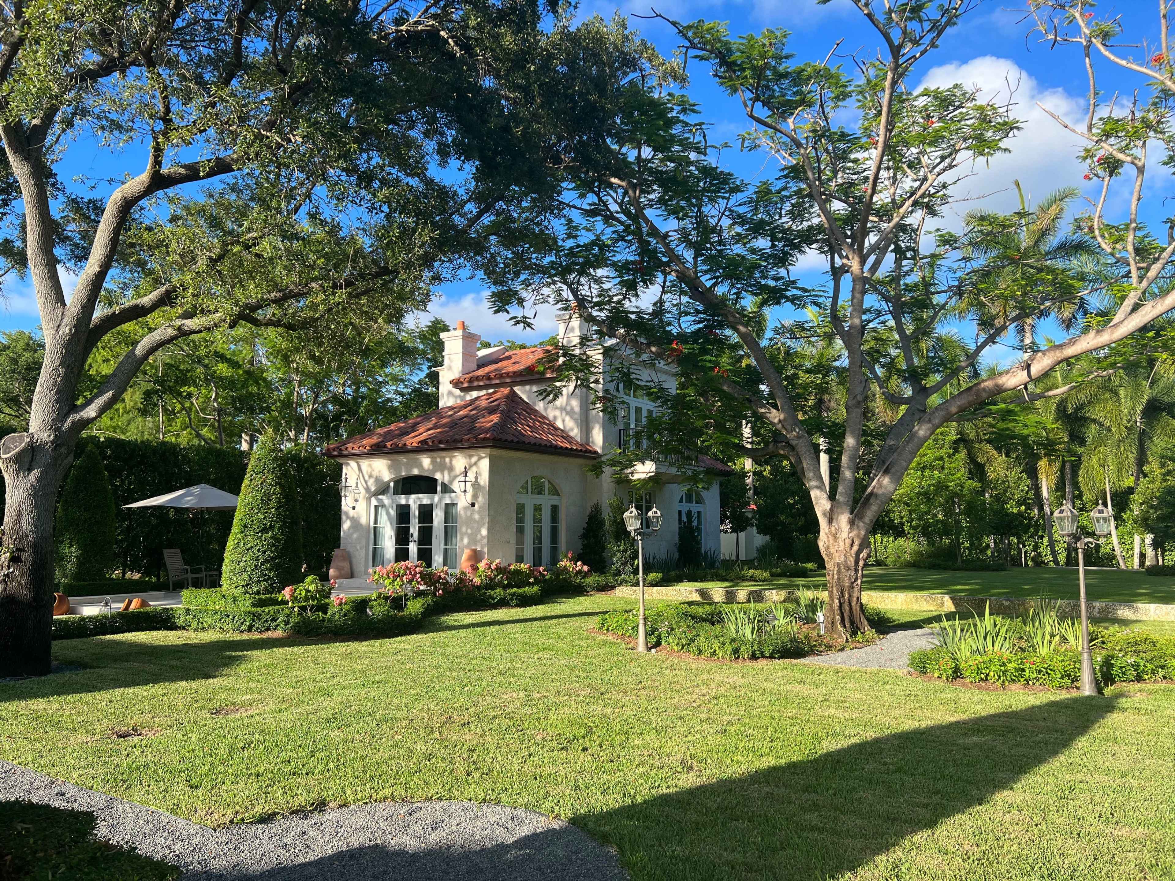 A large house with a red tile roof is surrounded by lush greenery and trees in a well-maintained garden.