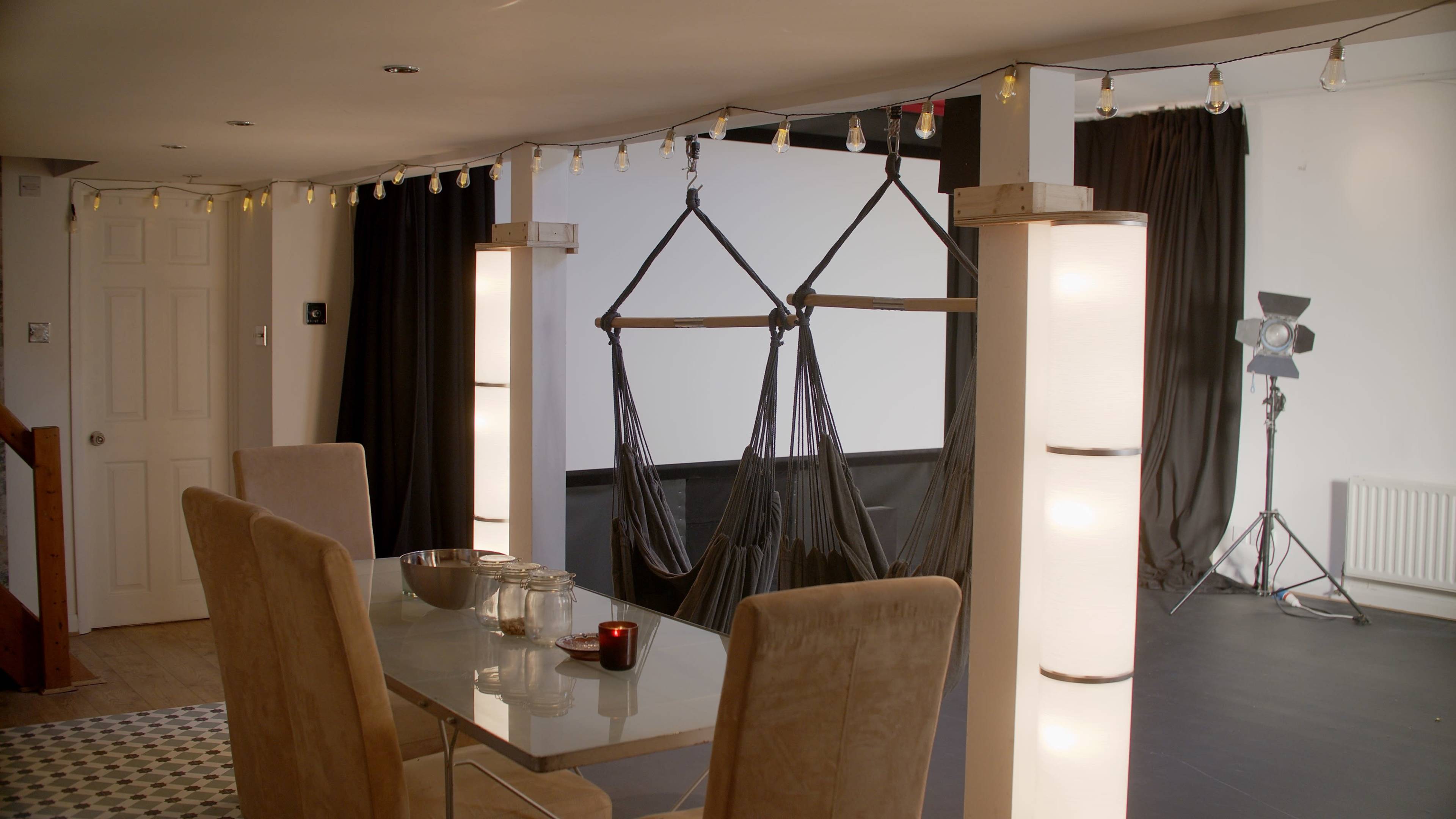 A modern indoor space featuring a dining table with chairs, decorative lighting, and a hammock suspended between two posts.
