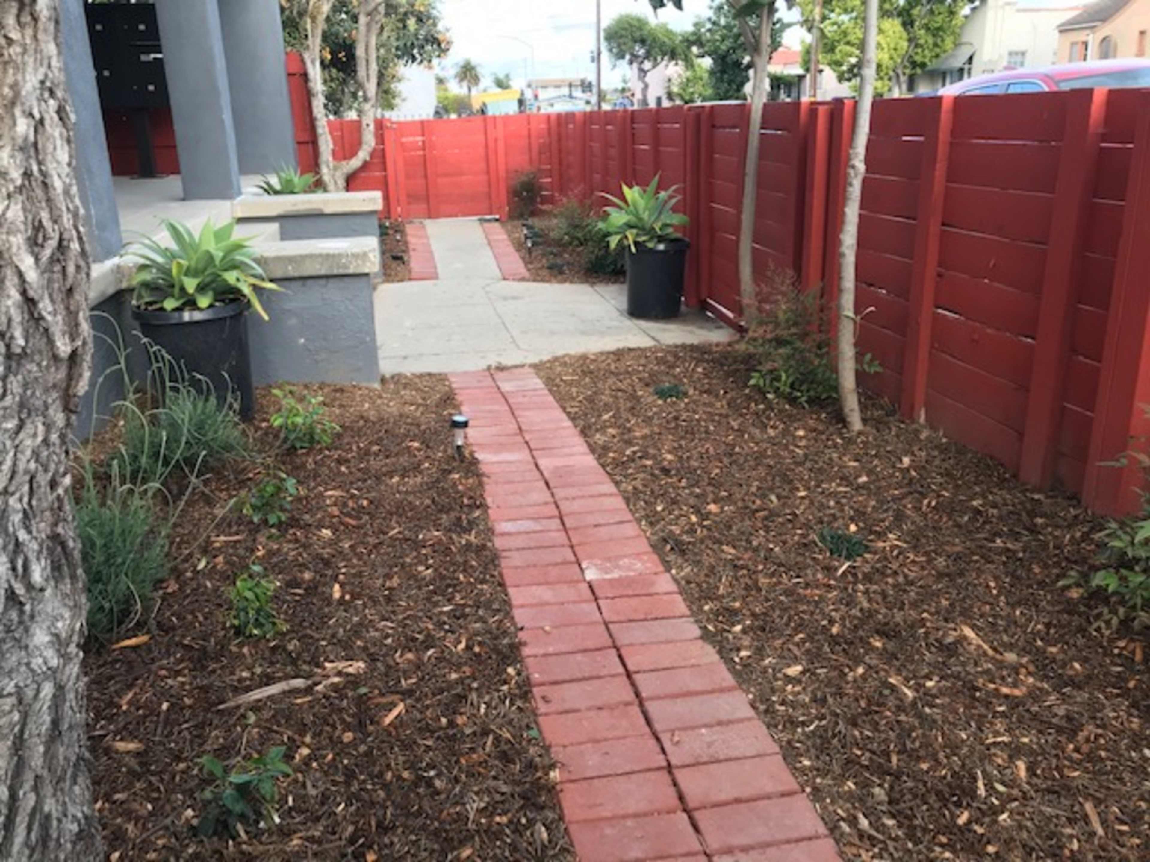 The image shows a landscaped walkway made of red bricks, bordered by mulch and small plants, leading between a red wooden fence and a tree.