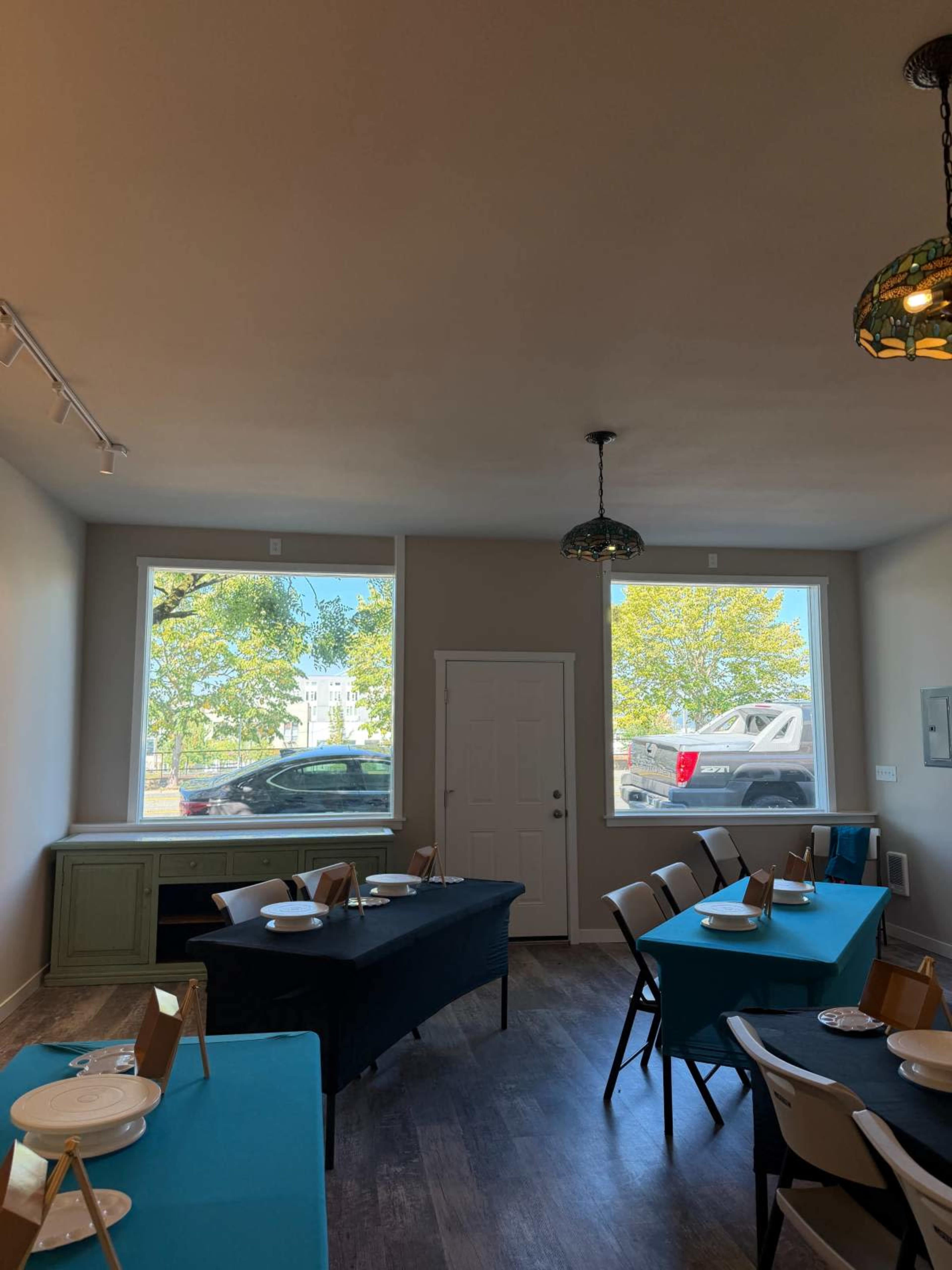 The image shows a bright dining space with tables set in black and turquoise linens, large windows providing natural light, and a view of a vehicle outside.