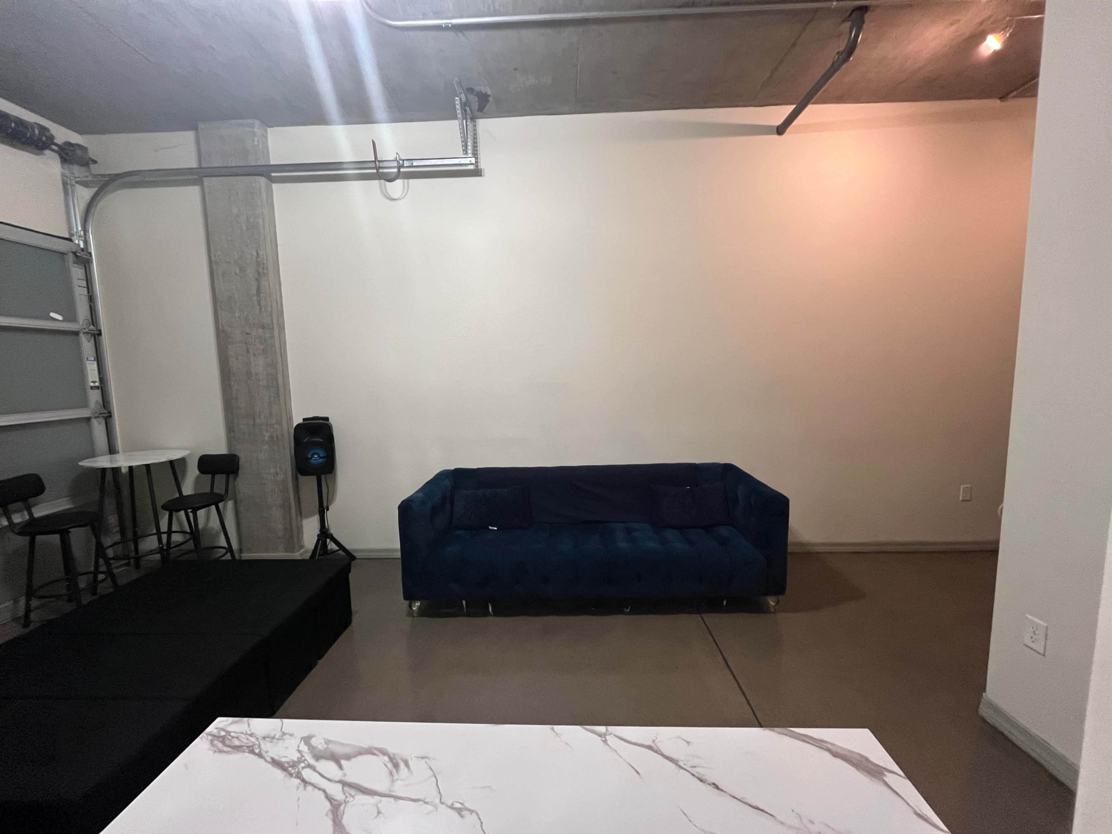 The image shows a room with a blue velvet couch, a small table with black chairs, and a stage area against a simple wall.