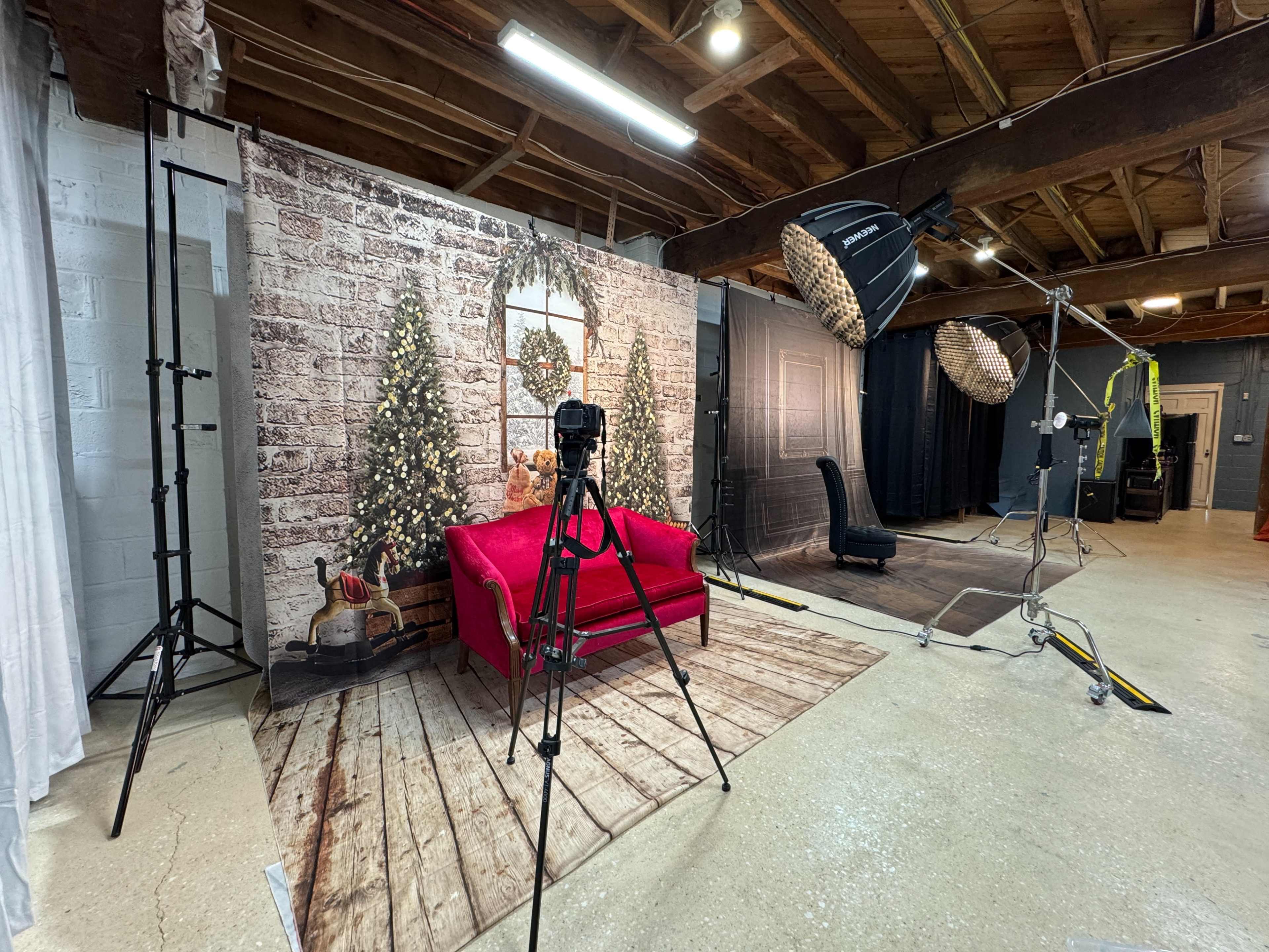 The image shows a photography studio with a brick-patterned backdrop featuring Christmas trees, a red couch, and various lighting equipment set up.