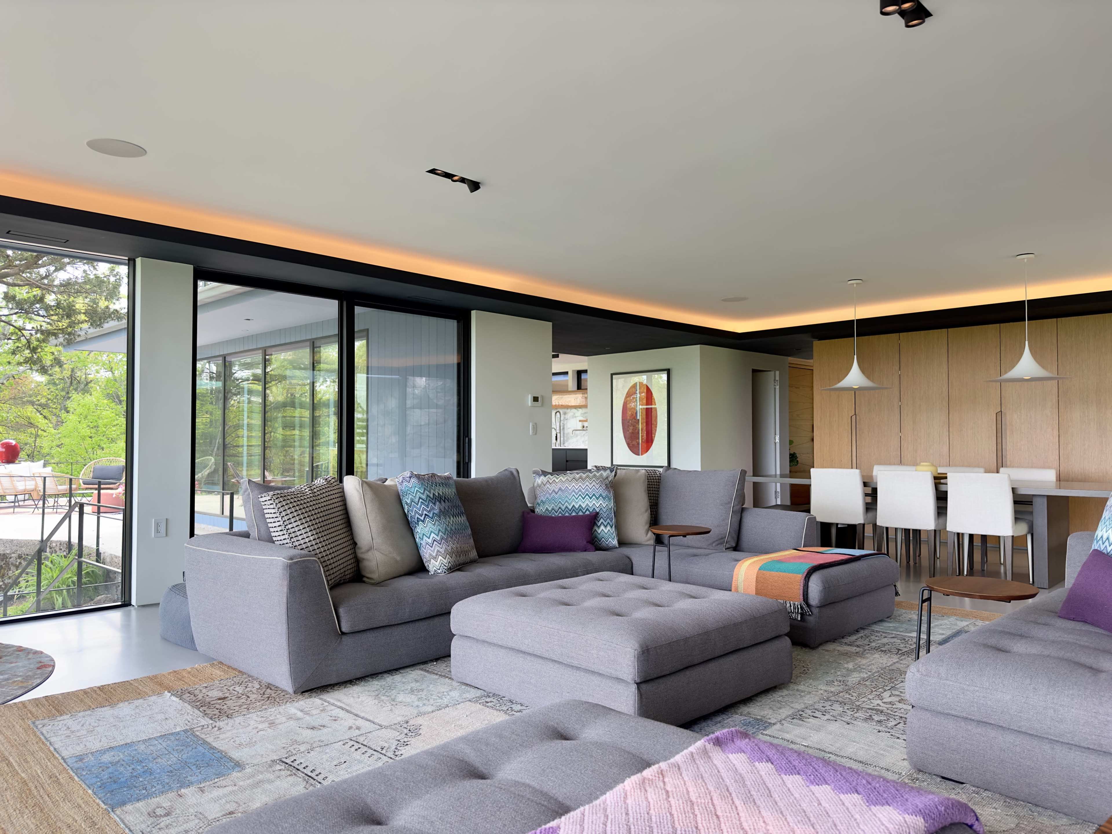 The image shows a modern living room with grey sectional sofas, a large area rug, and a dining area visible in the background.