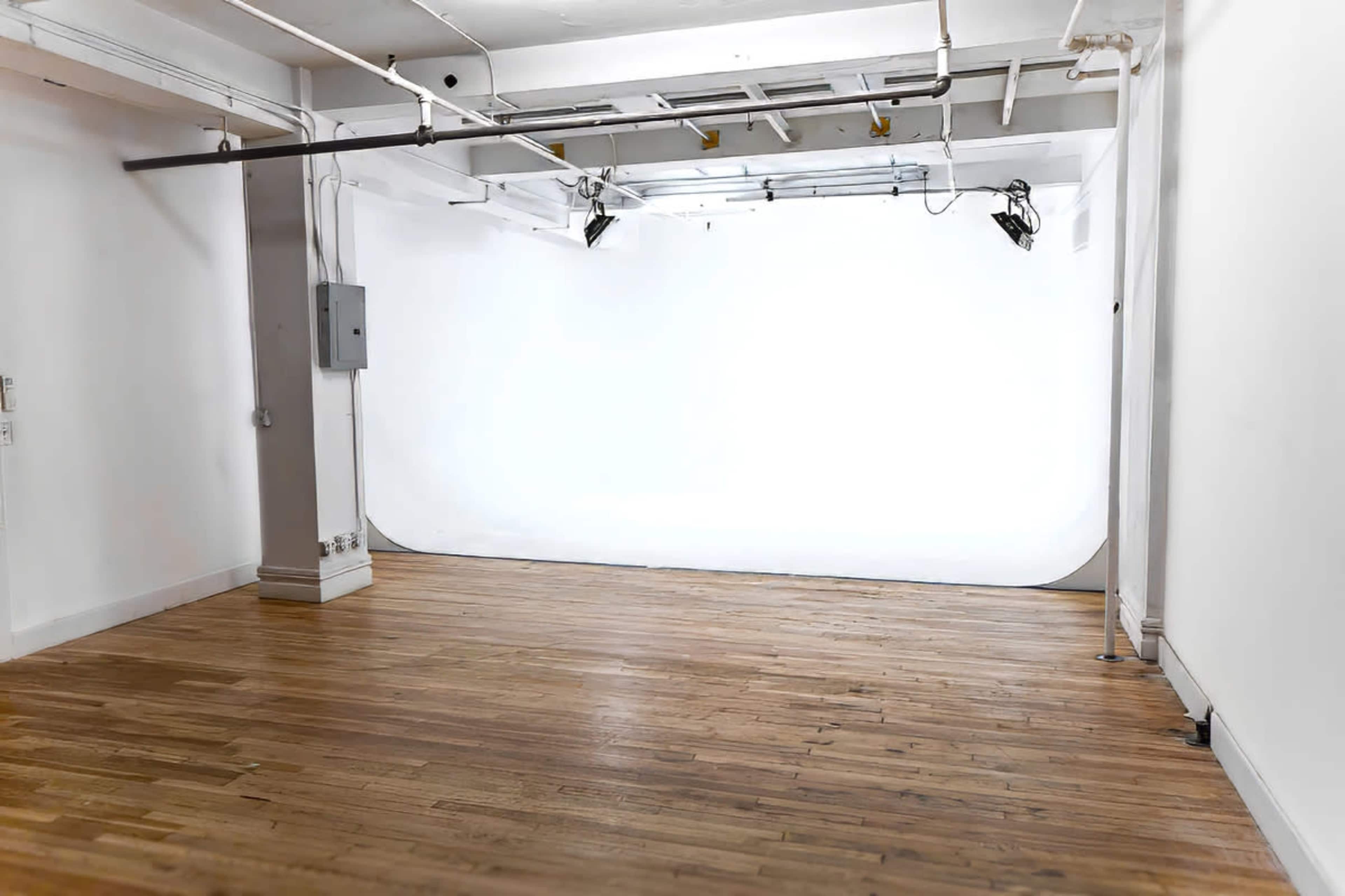 The image shows an empty interior studio space with wooden flooring and a white backdrop at the far end.
