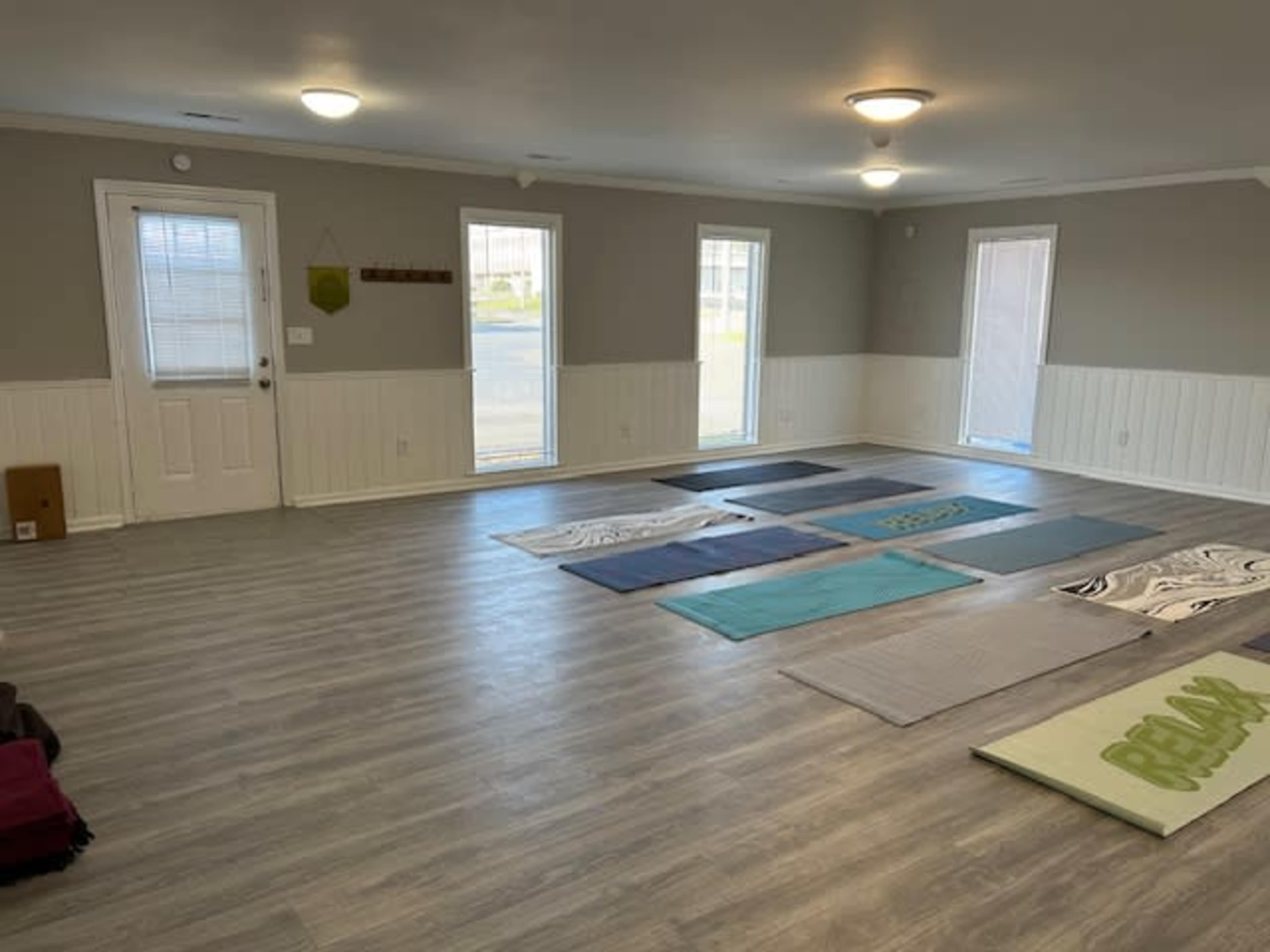 A spacious room with gray walls and light wood flooring features several exercise mats arranged on the floor and large windows letting in natural light.