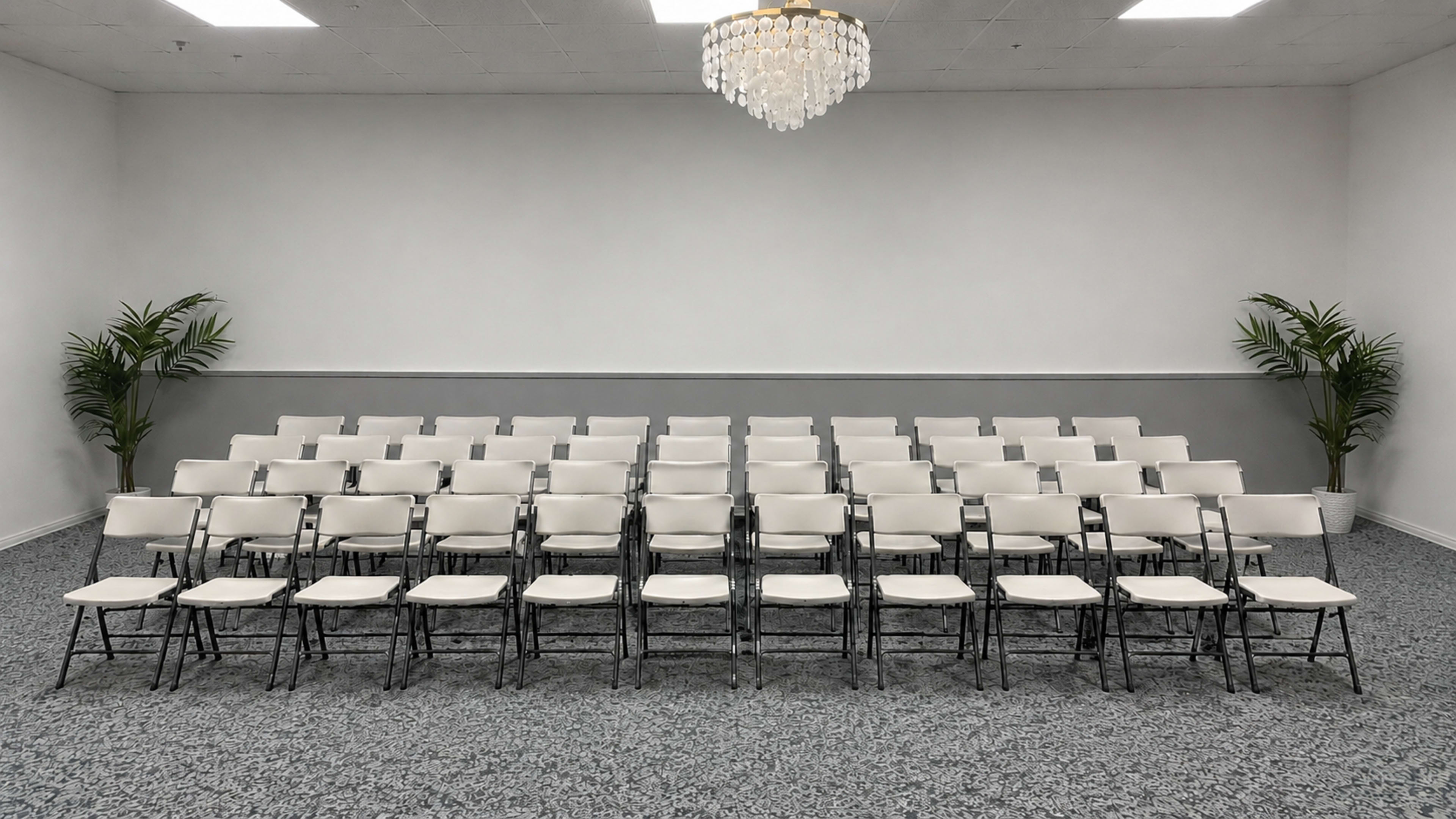 The image shows a row of white folding chairs arranged neatly in a spacious indoor area with a chandelier and potted plants at either side.