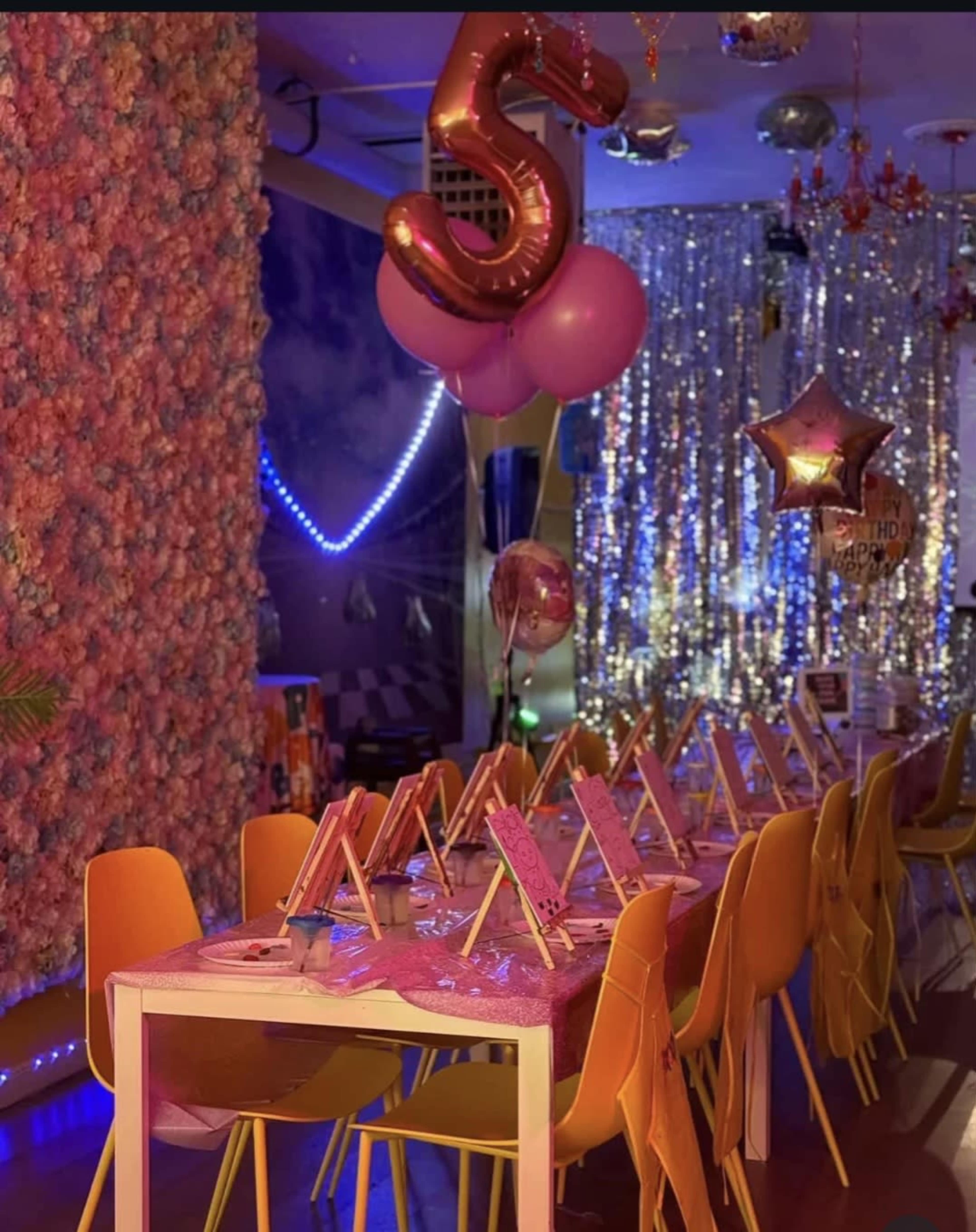 The image shows a festive party setup with a table decorated in pink, yellow chairs, and balloons, set against a backdrop of shimmering decorations.
