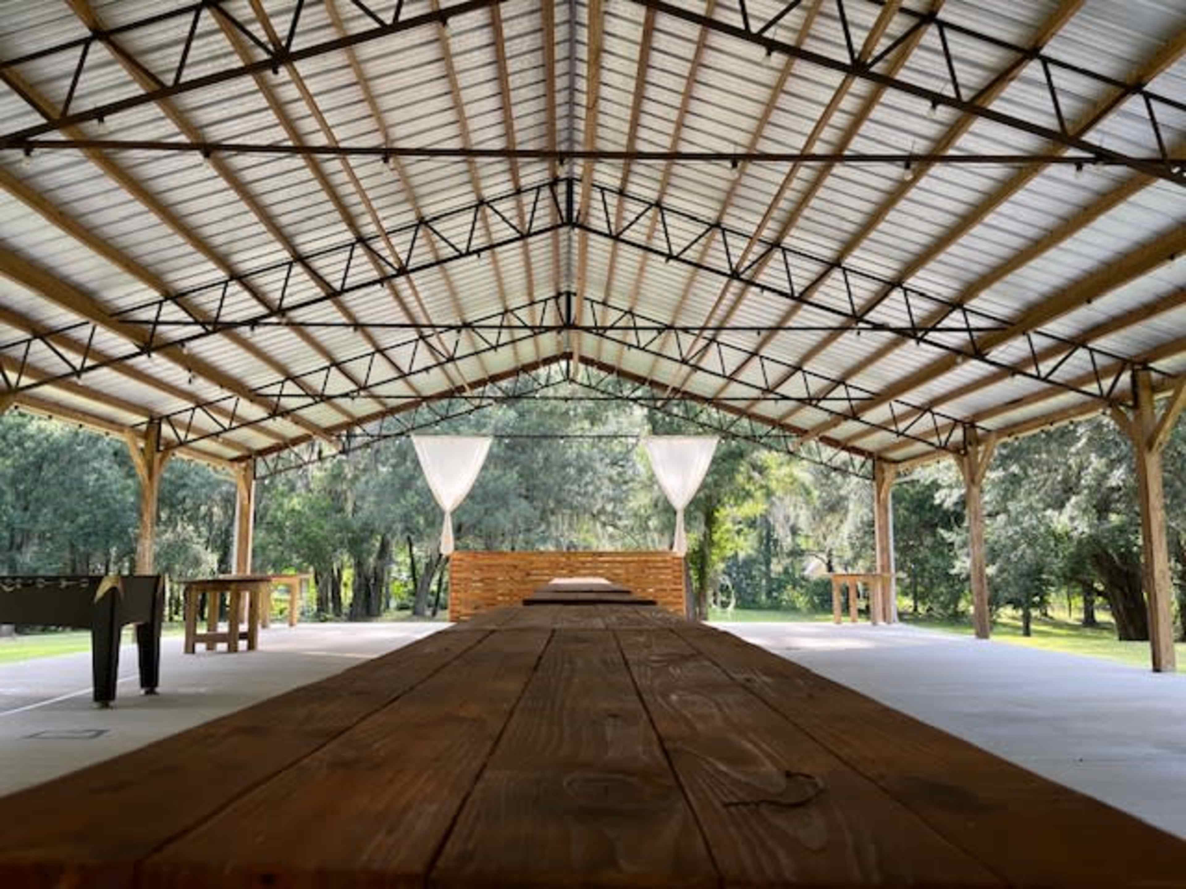 A spacious, open pavilion with wooden beams and a long table set in a green outdoor area.