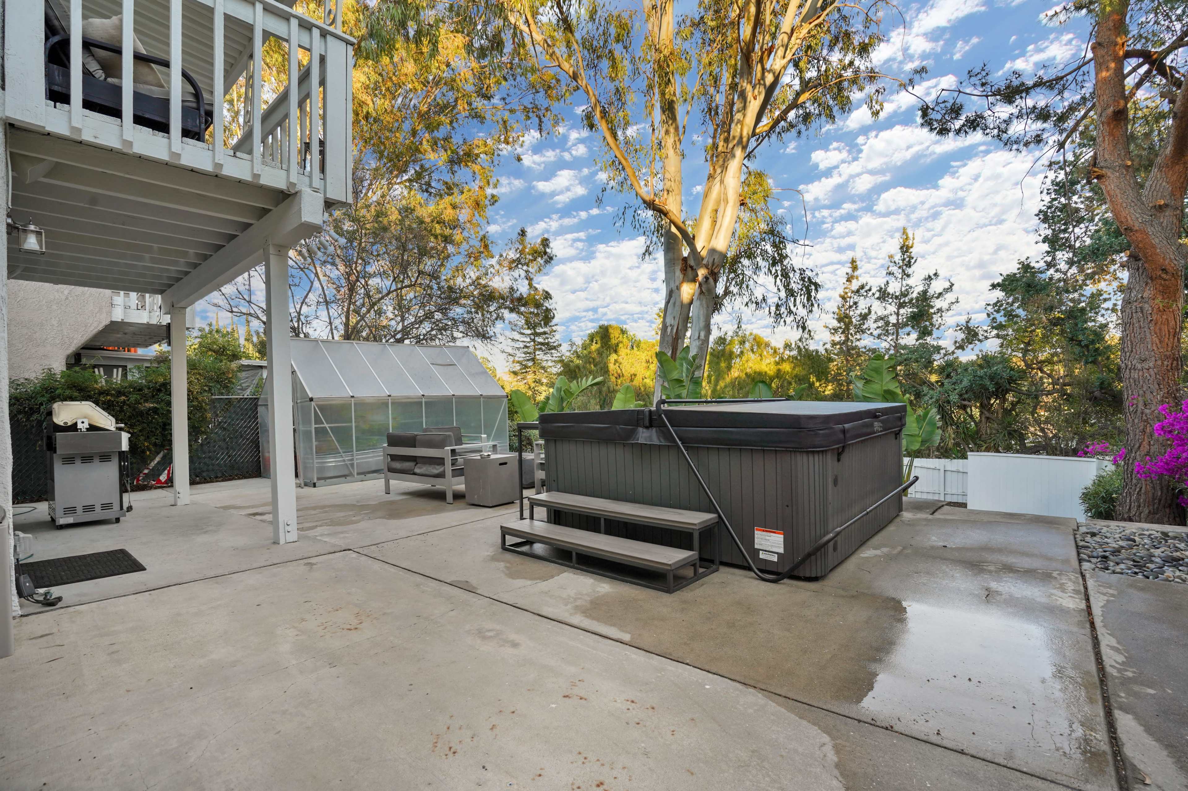 The image shows a backyard patio featuring a hot tub, outdoor seating area, and a greenhouse surrounded by trees and plants.