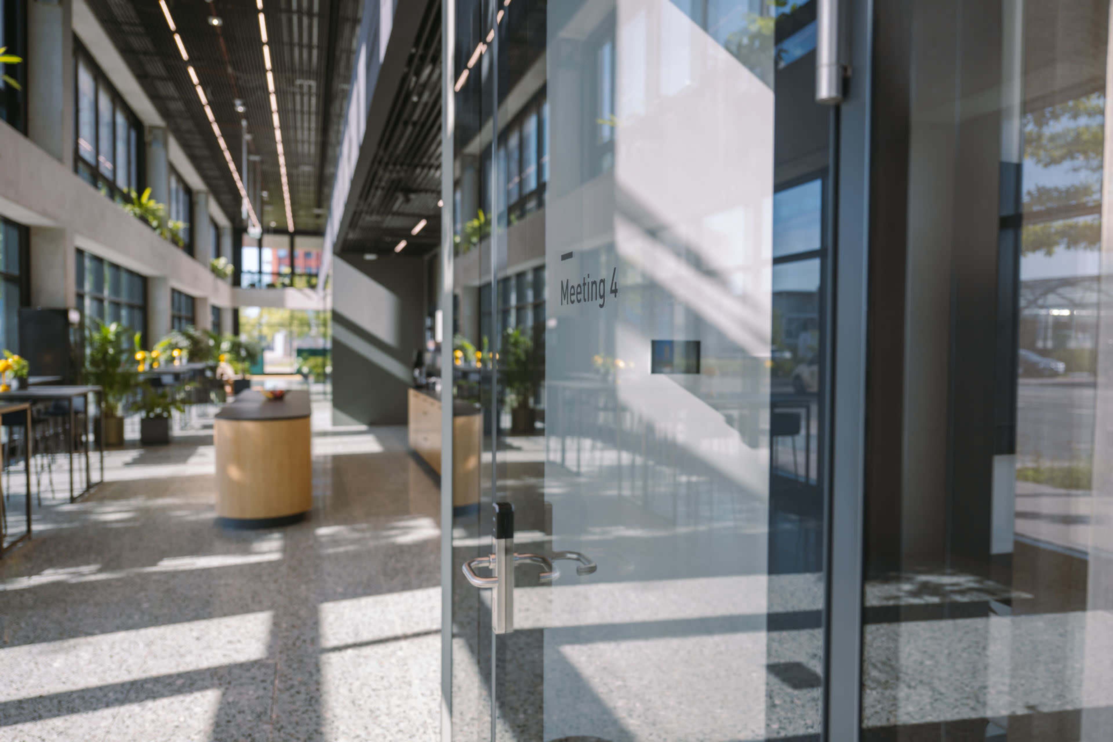 A glass door labeled "Meeting 4" opens into a spacious, well-lit office area with plants and modern furniture.
